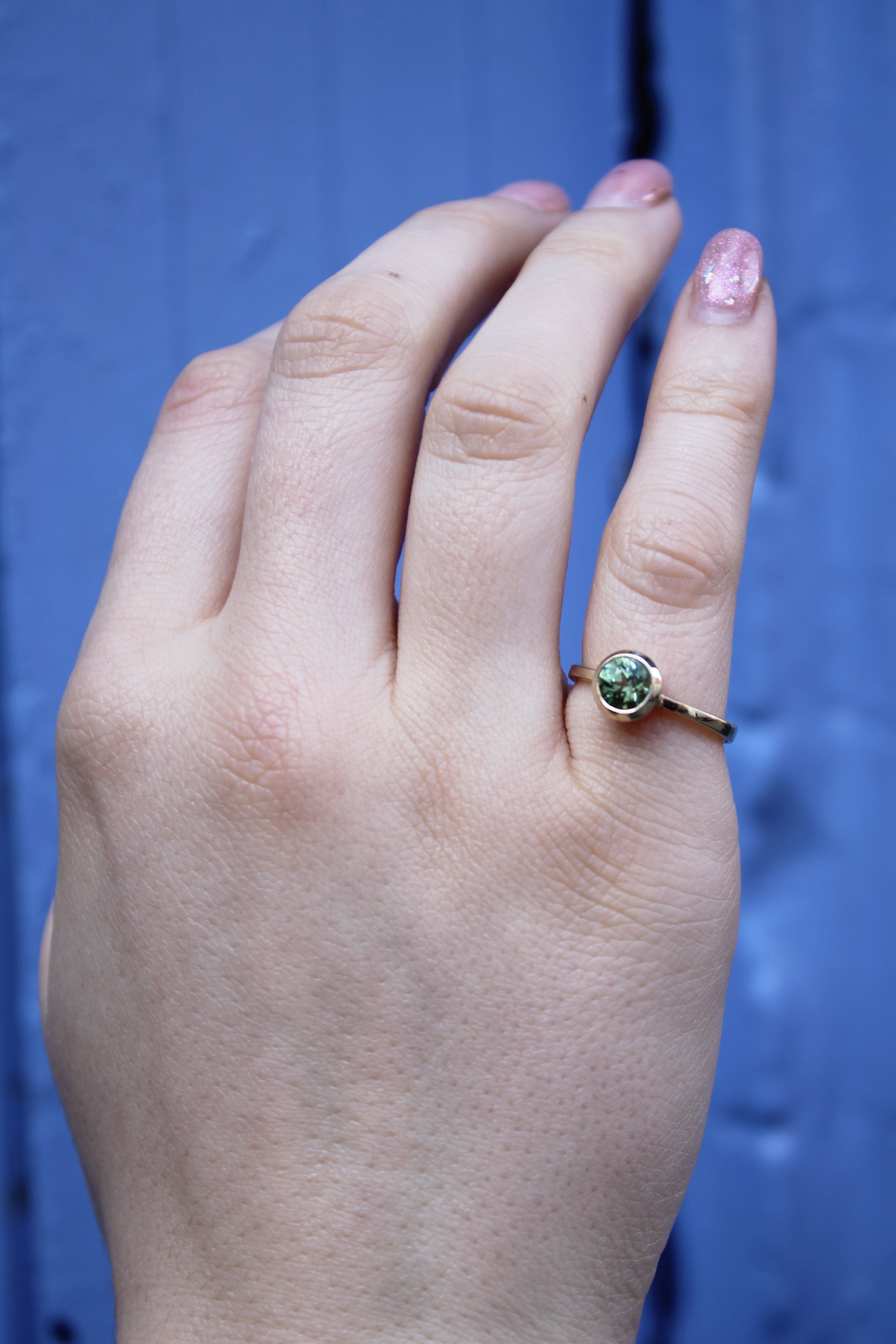 A hand with light skin and pink glittery nails wears the Madeleine Fine Jewellery Viridian Dawn 9ct yellow gold ring, featuring a round green gemstone, shown against a blue background.