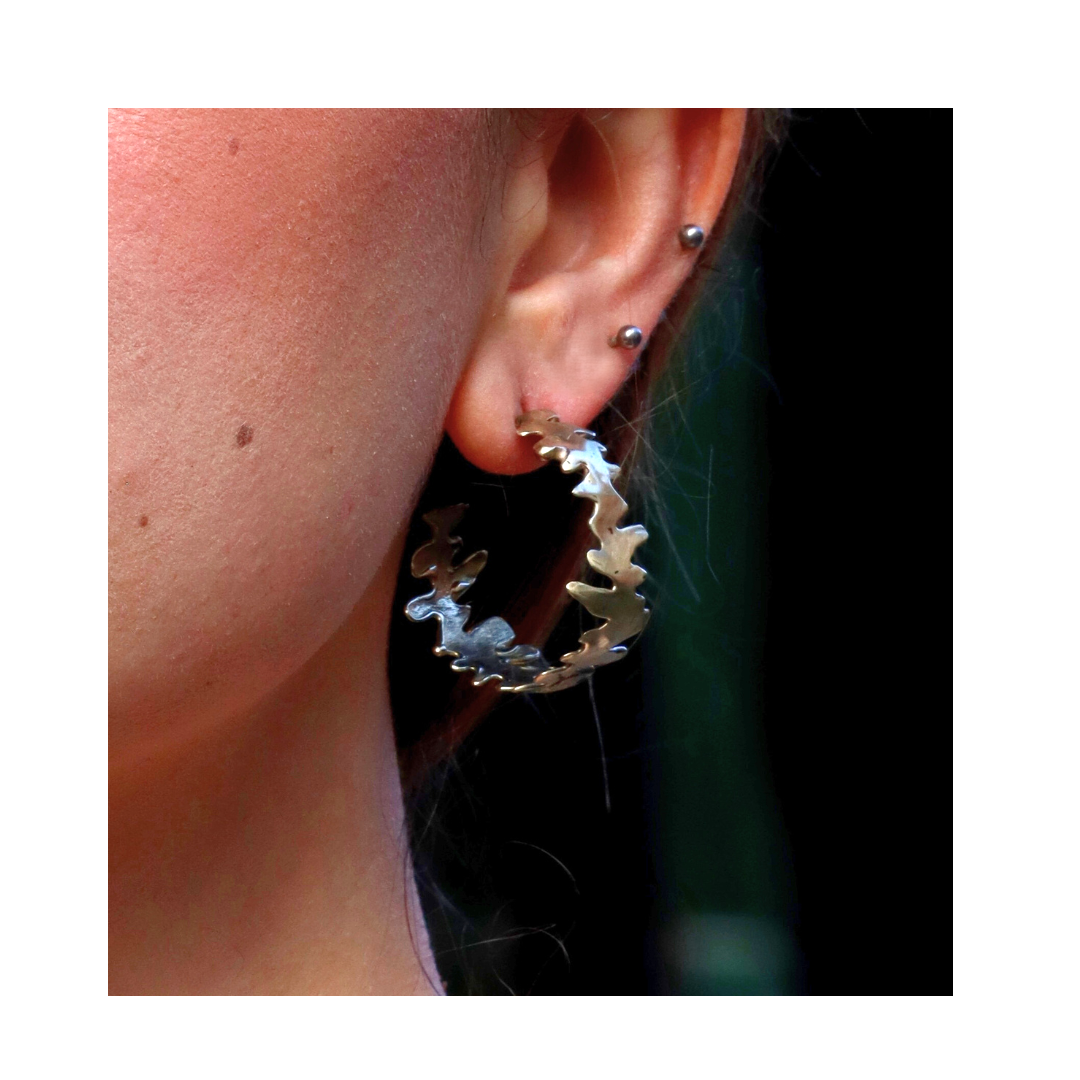 Close-up of a person’s ear adorned with the Madeleine Blaine Lava Hoops—a large, jagged silver hoop earring—paired with two small silver studs above it. The dark, blurred background accentuates the sculptural, hand-carved jewelry from the Lava Hoops colle
