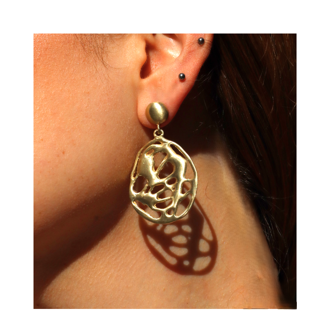 Close-up of an ear adorned with three small studs and a large, round, abstract gold earring—reminiscent of Madeleine Blaine’s Molten Drop Earrings—casting a shadow on the person’s neck. The bold, hand-carved Molten Drop Earring shines brilliantly against