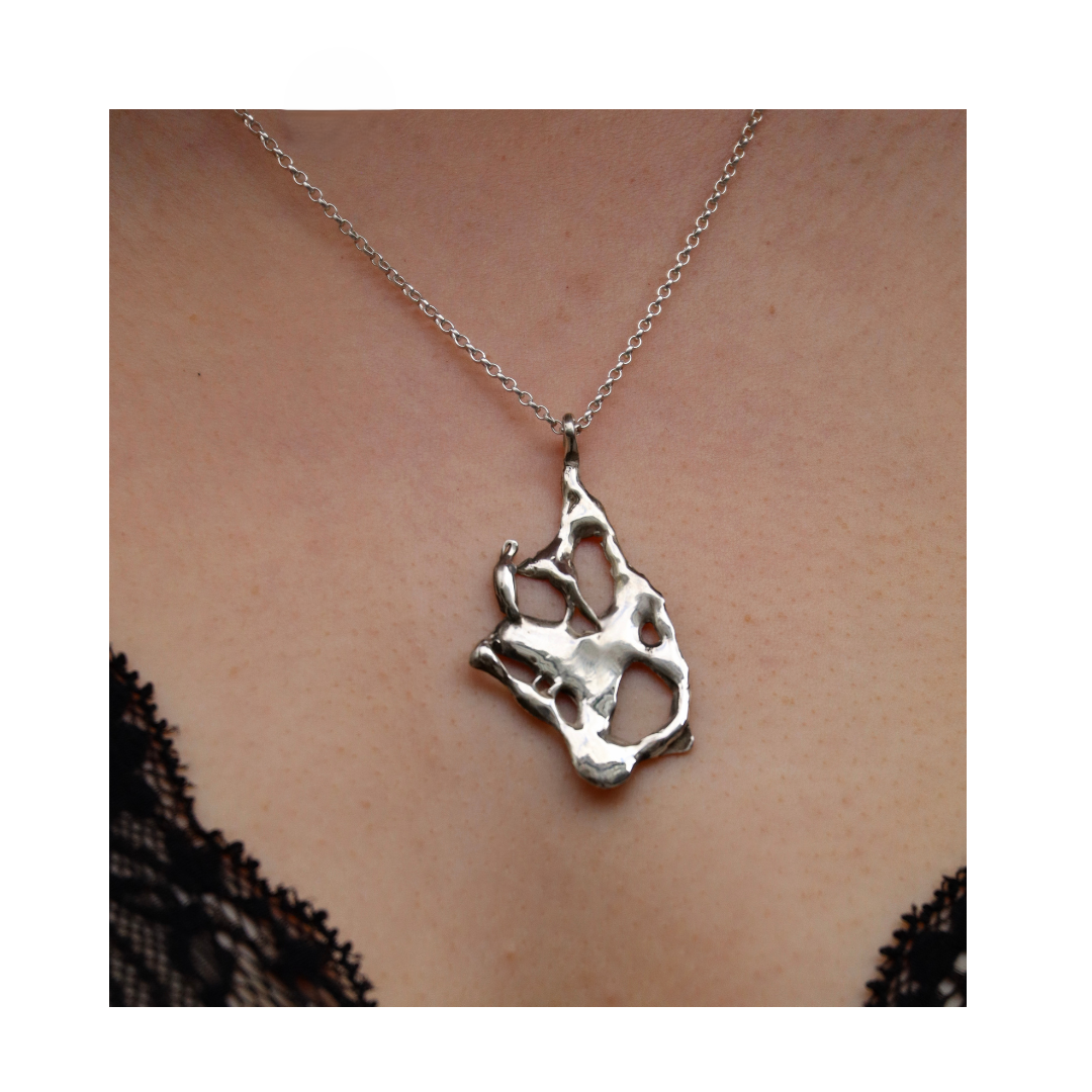 A close-up shows a person wearing the Molten Necklace by Madeleine Sculptural Jewellery—a silver abstract pendant on a chain—contrasted against skin and black lace, highlighting the elegance of sustainable jewellery.