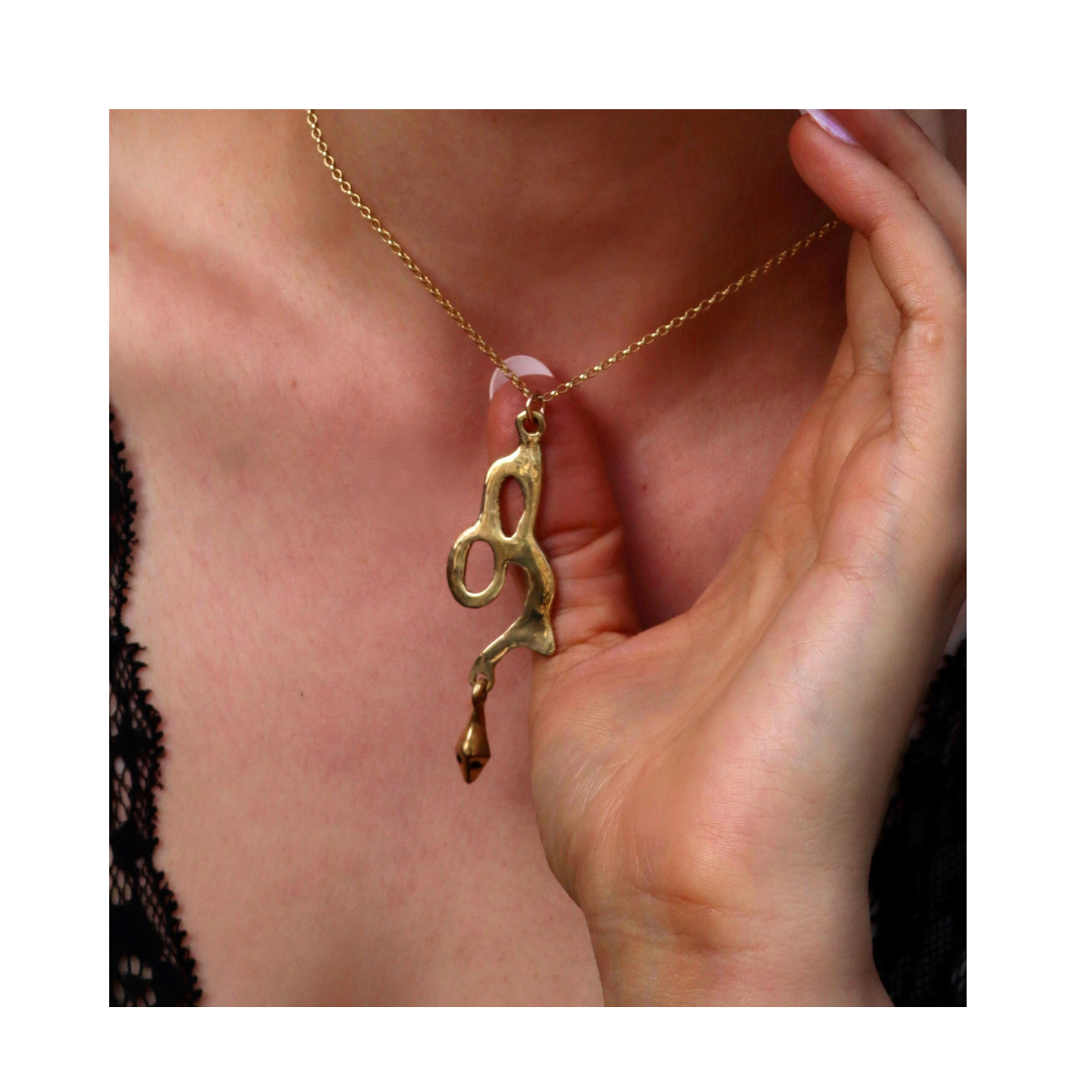 A hand holds the Serpent Pendant by Madeleine Sculptural Jewellery, a gold abstract-shaped necklace crafted from recycled brass, shown against a black lace top.