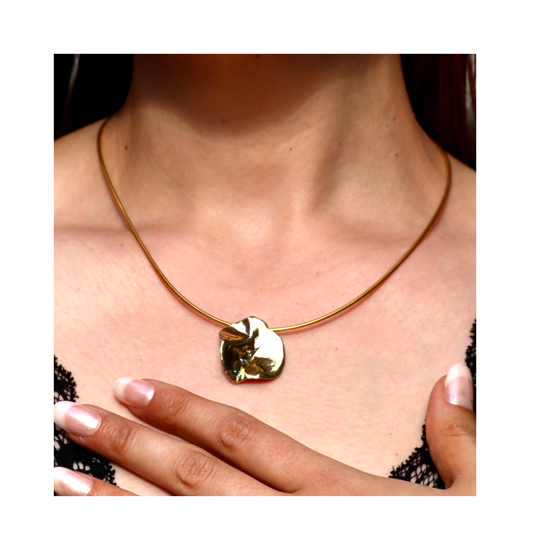 A close-up of a person wearing the Tide Necklace by Madeleine Sculptural Jewellery—a gold, round, textured pendant. Their French-manicured hand rests on their chest, framed by organic black lace.