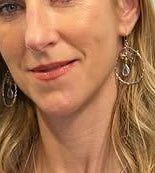 A close-up shows a woman with long blonde hair, soft makeup, and a gentle smile, wearing Watermelon Tropical's Wildlife Large Bee Hoop Drops in various gemstones. Only the lower half of her face and part of her earrings are visible.