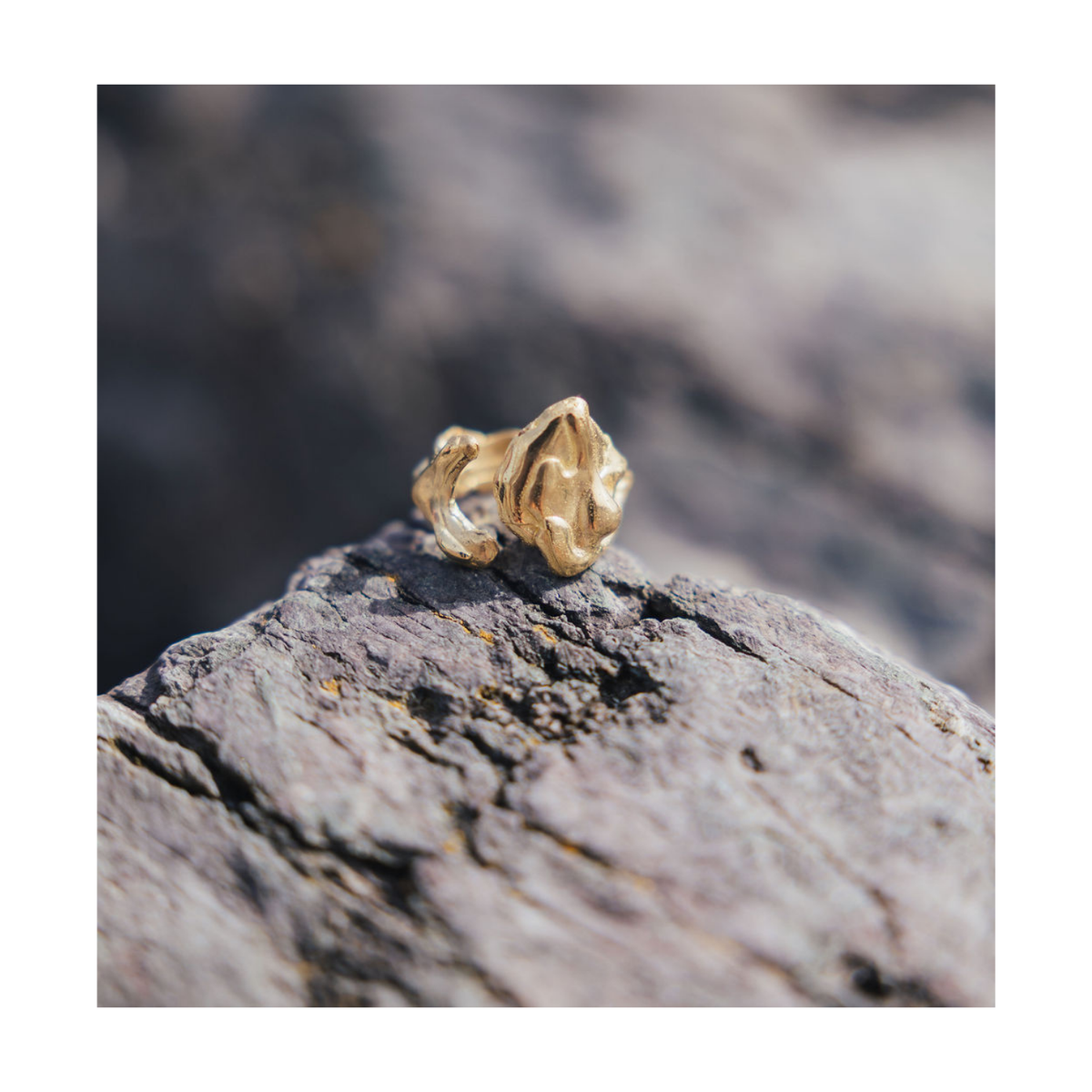 A hand-carved gold Blaze Ring by Madeleine Blaine, featuring an organic, textured design, is showcased on a rough gray rock surface, reflecting the artistry of Dublin craftsmanship.