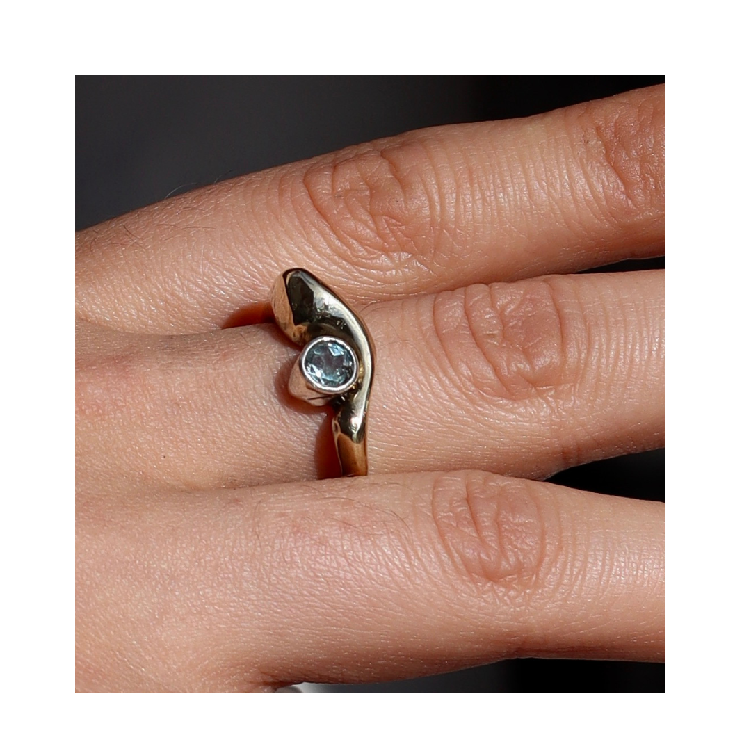 A close-up of a hand wearing the Bough Ring Blue Topaz by Madeleine Sculptural Jewellery, a handmade gold ring with a wavy design and a single round blue topaz gemstone set in the center, worn on the middle finger.