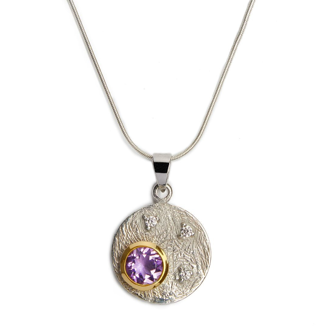 The Celeste Necklace by Gallardo & Blaine Designs features a sterling silver chain with a round textured pendant, set with a central purple gemstone in gold and accented by three sparkling cubic zirconia stones.