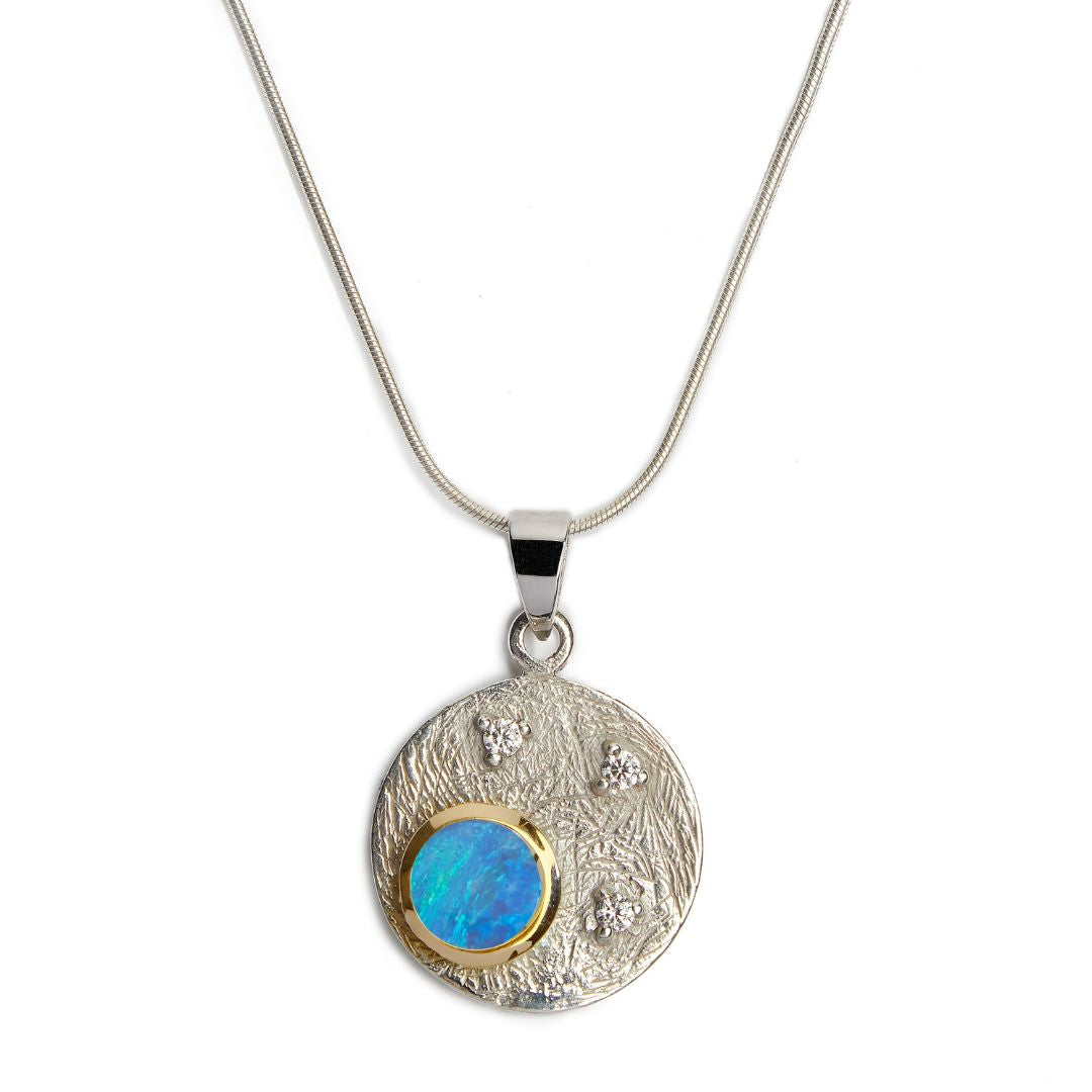 The Celeste Necklace by Gallardo & Blaine Designs features a sterling silver chain with a round, textured pendant set with a blue-green opal in gold and accented by four cubic zirconia stones.