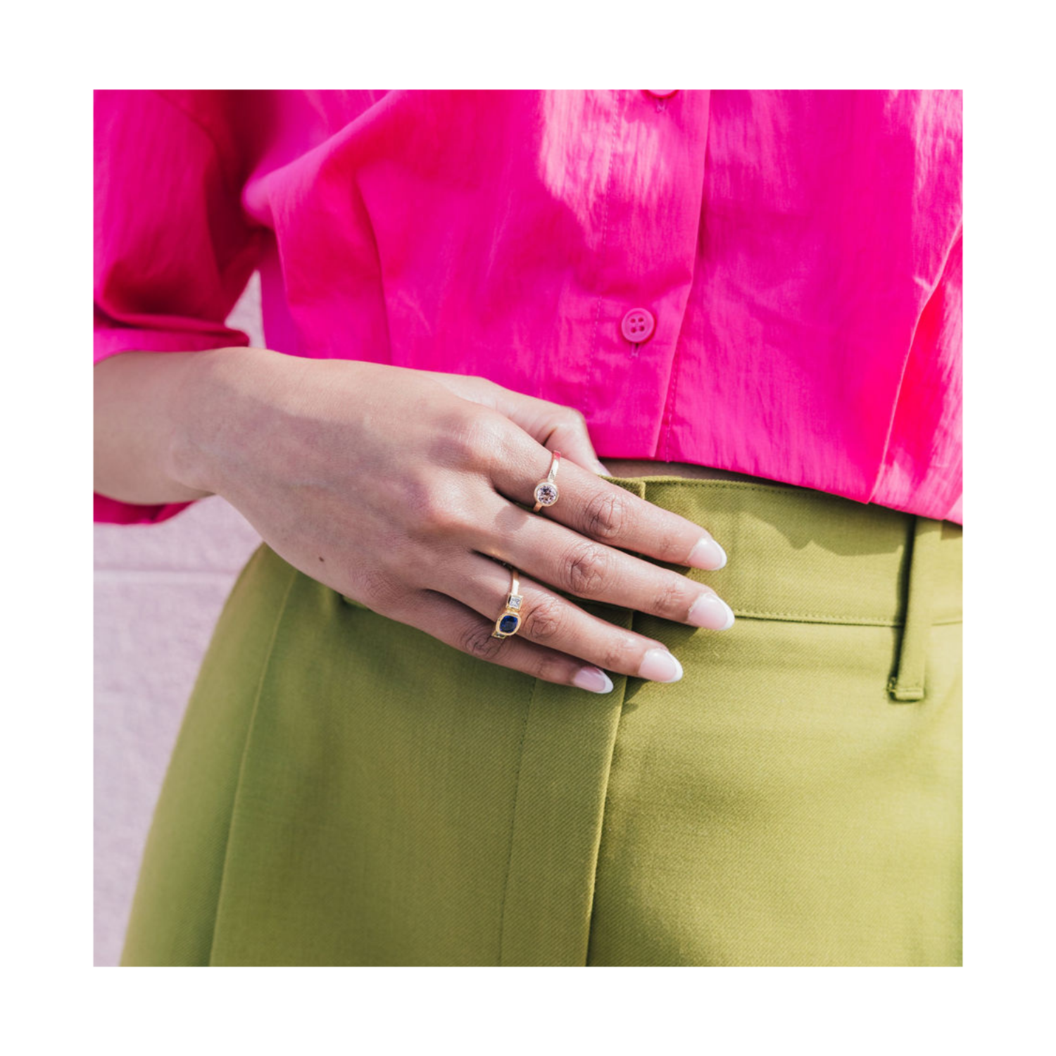 A close-up of a person's hand resting on their lap, wearing two silver rings: one with a small clear heart-shaped stone and another with a dark oval stone. The person is wearing a bright pink button-up shirt and olive green trousers.