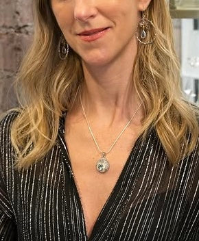 A woman with blonde wavy hair wears a black blouse with white stripes, drop earrings, and the Gallardo & Blaine Designs Celeste Necklace in Silver with gemstone, smiling softly.