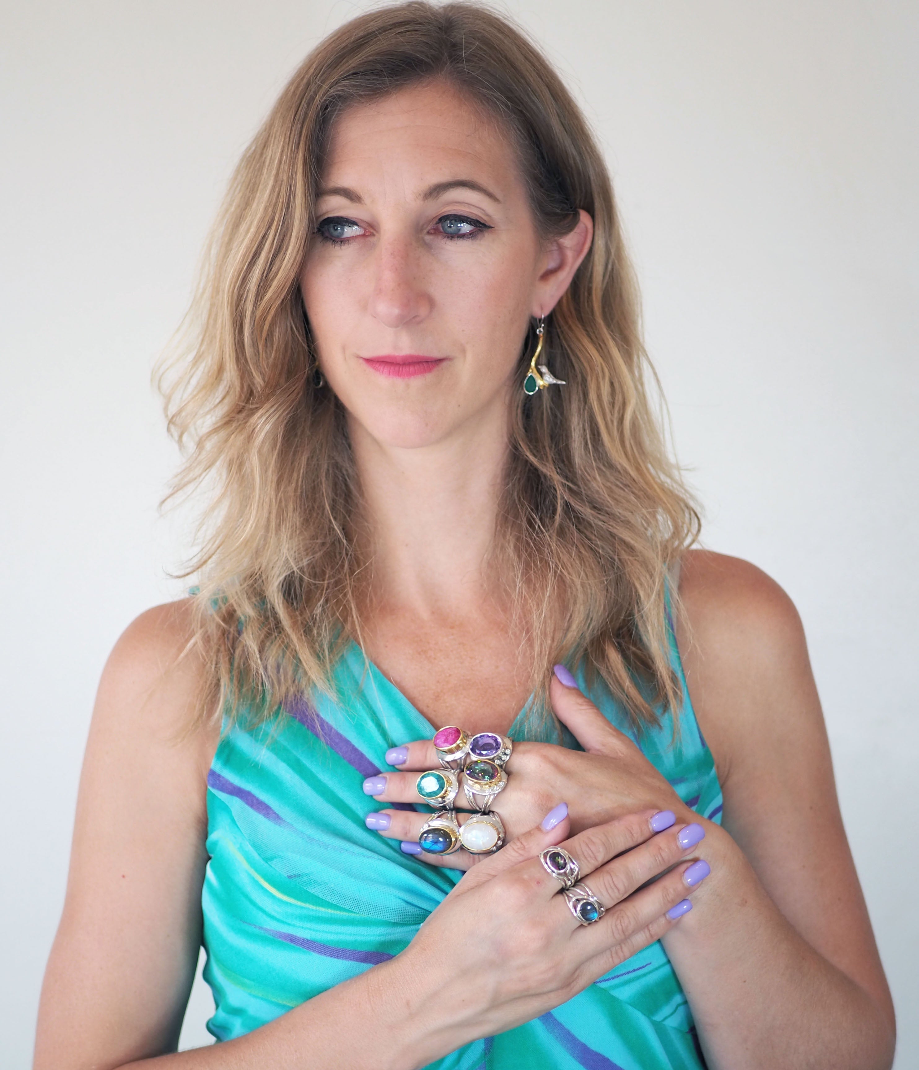 A woman with wavy blonde hair, wearing a blue-green dress and lavender nails, poses with her hands on her chest to showcase the Eyetelia Ring in various gemstones by Gallardo & Blaine Designs. She looks slightly to the side.