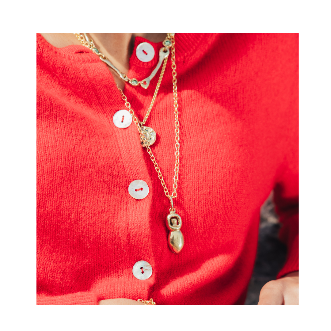 A person wearing a bright red cardigan with white buttons, layered gold necklaces, and the Danu Pendant by Madeleine Sculptural Jewellery, shaped like an abstract figure.