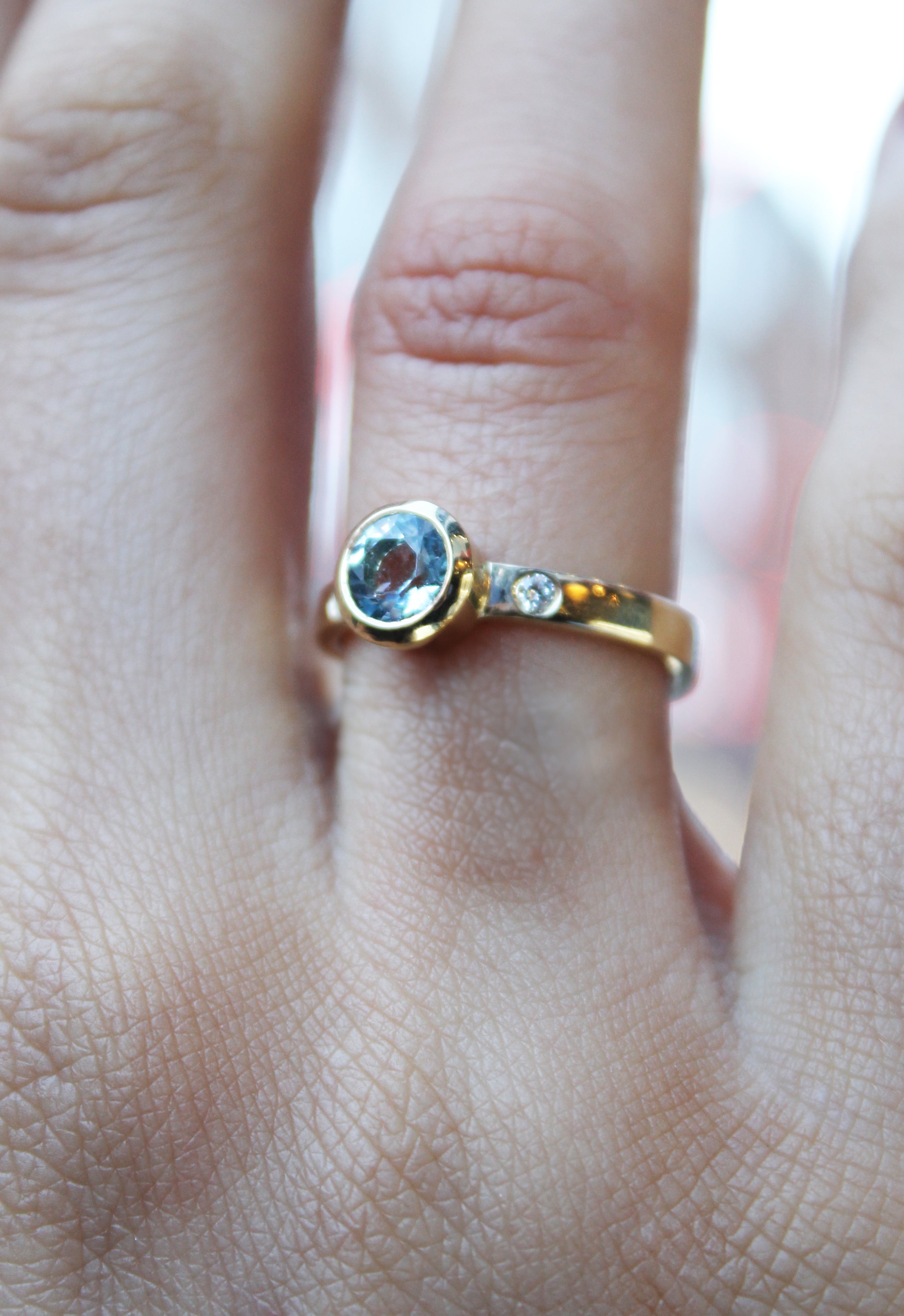 A hand wears the Timeless Cascade 9ct Yellow Ring by Madeleine Fine Jewellery, featuring a large oval aquamarine and a small lab grown diamond, shown on the ring finger.