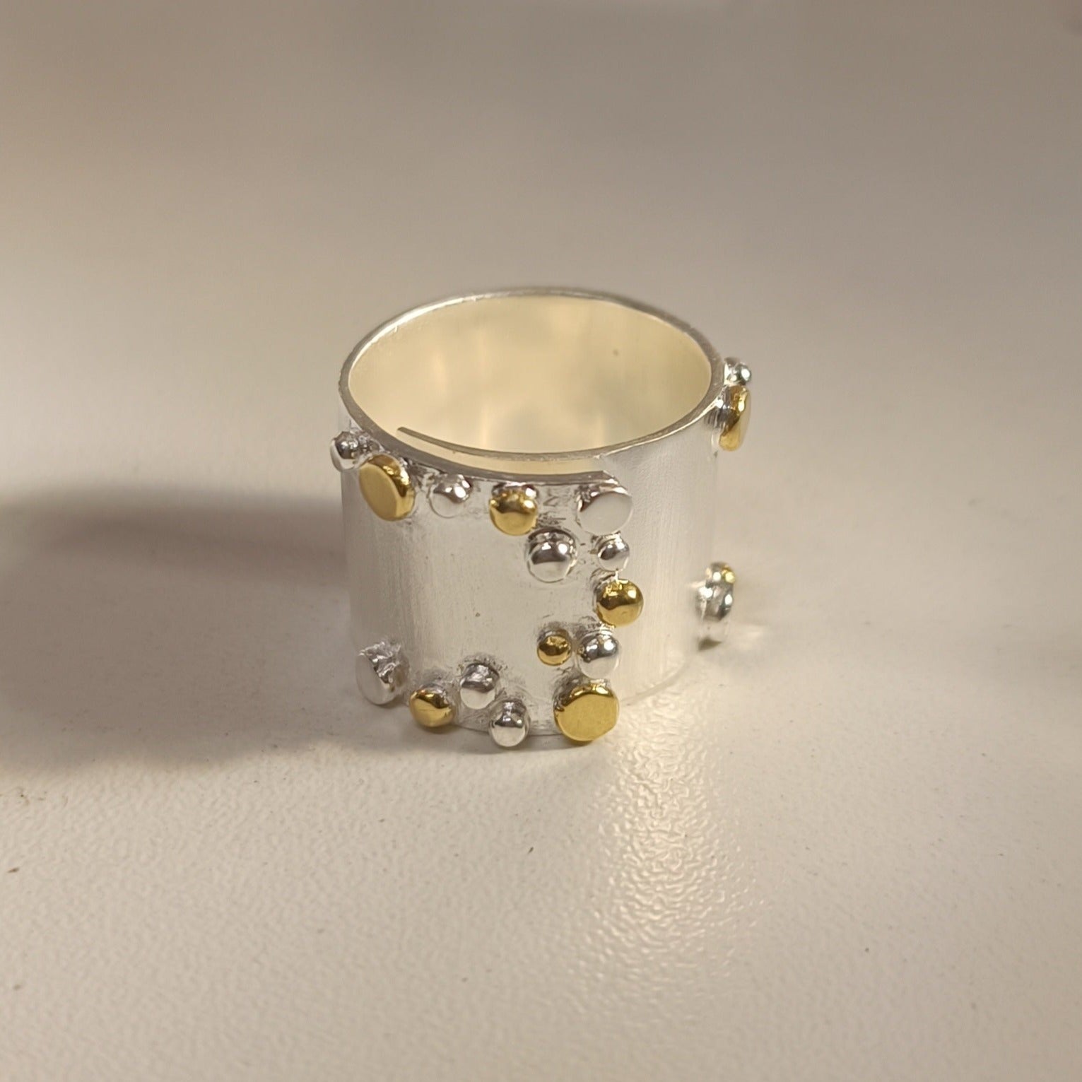 The Adjustable Iona Silver & Gold Ring by Gallardo & Blaine Designs is a wide, matte-finished 925 sterling silver piece with an abstract design featuring small silver and gold spheres on its surface, set against a simple light background.