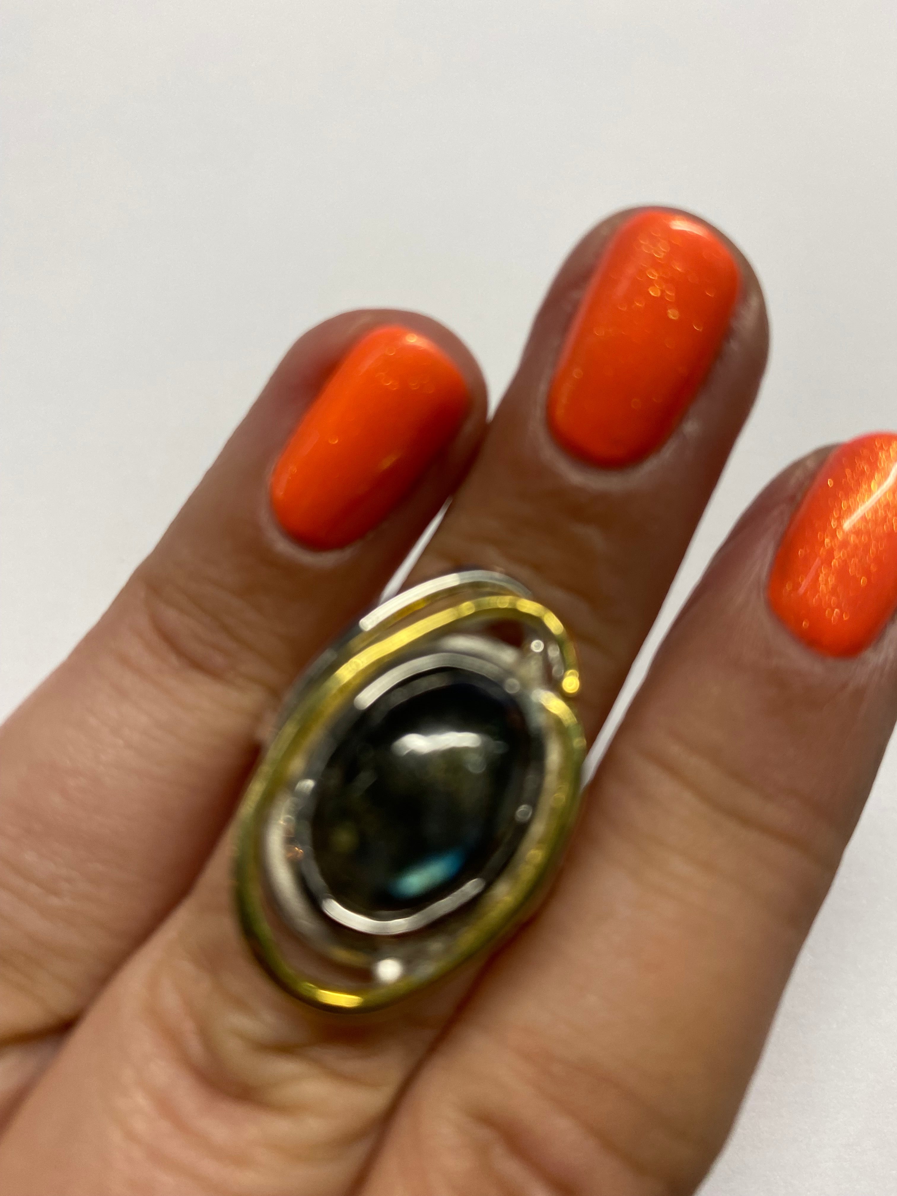 A close-up of a hand with brightly painted orange nails, showcasing a large, ornate silver ring. The gemstone is an oval-shaped, dark stone encased in intricate gold plate metalwork. The background is plain and out of focus. The product being featured is 