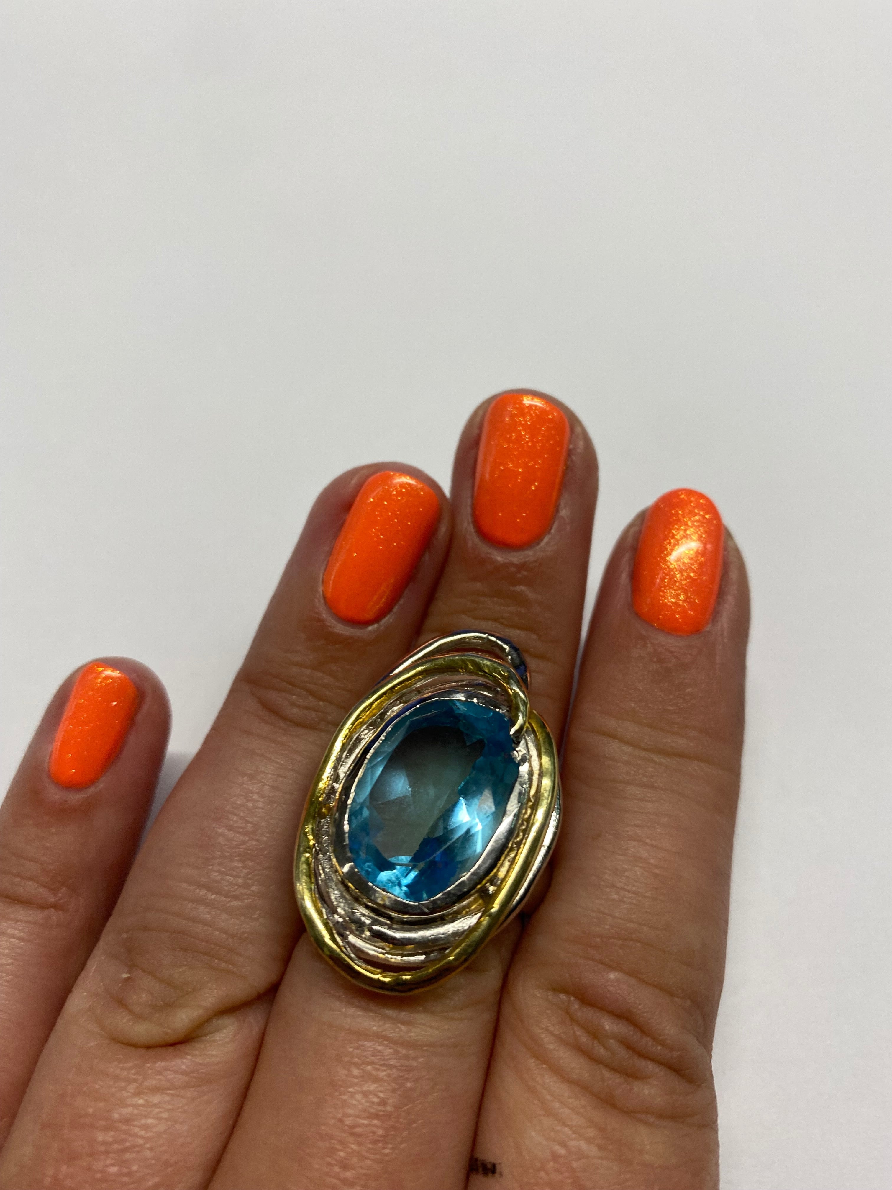 A hand with bright orange, glittery nail polish is shown against a white background. The ring finger flaunts a large gold ring with an oval-shaped blue gemstone, reminiscent of a Cosmos Ring Large in Various Stones by Gallardo & Blaine Designs.