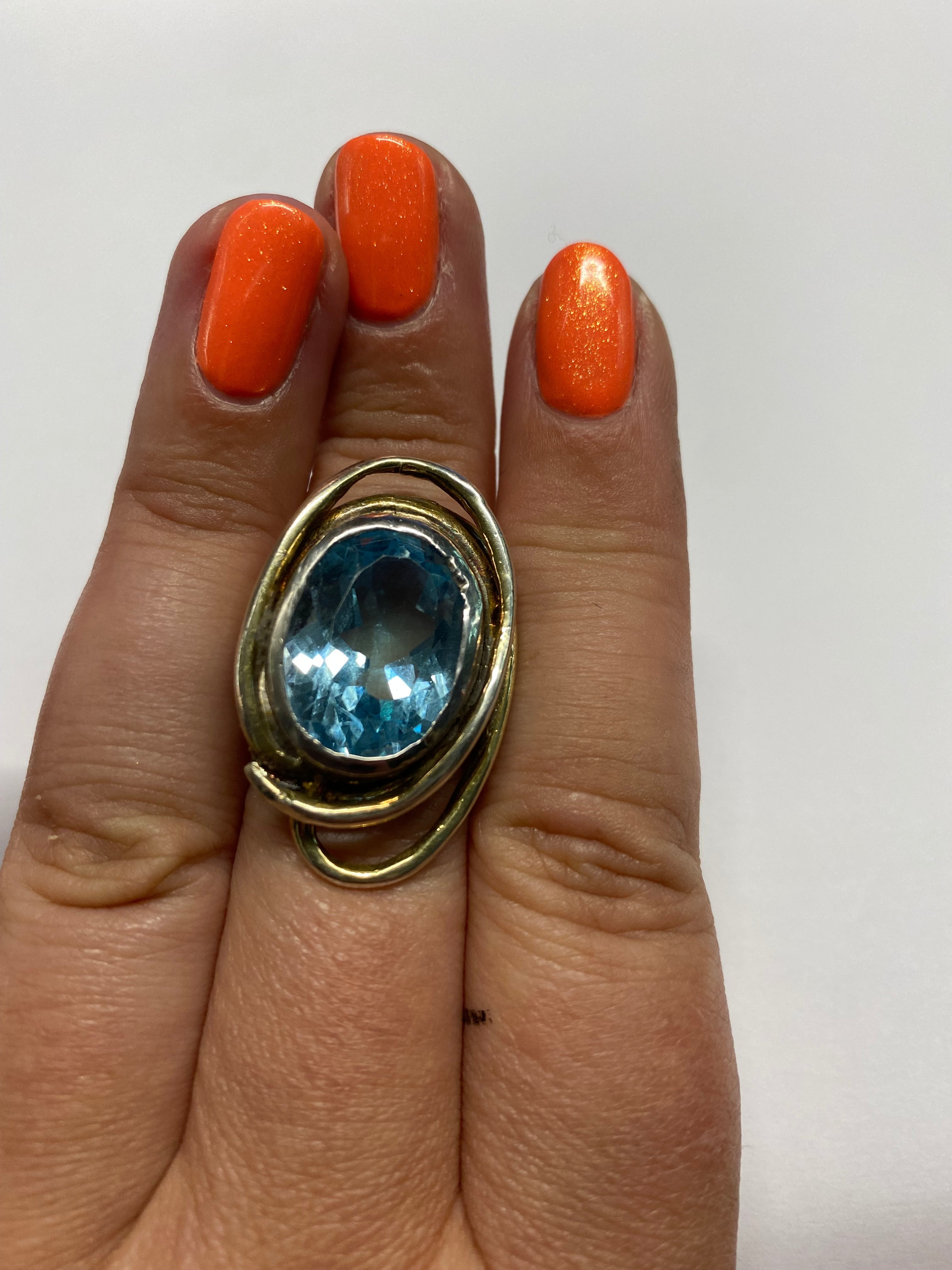 A hand with bright orange painted nails is wearing a Gallardo & Blaine Designs Cosmos Ring Large in Various Stones, featuring a large, oval-shaped blue gemstone. The ring's band is a unique twisted design, and the gemstone is prominently showcased in the 