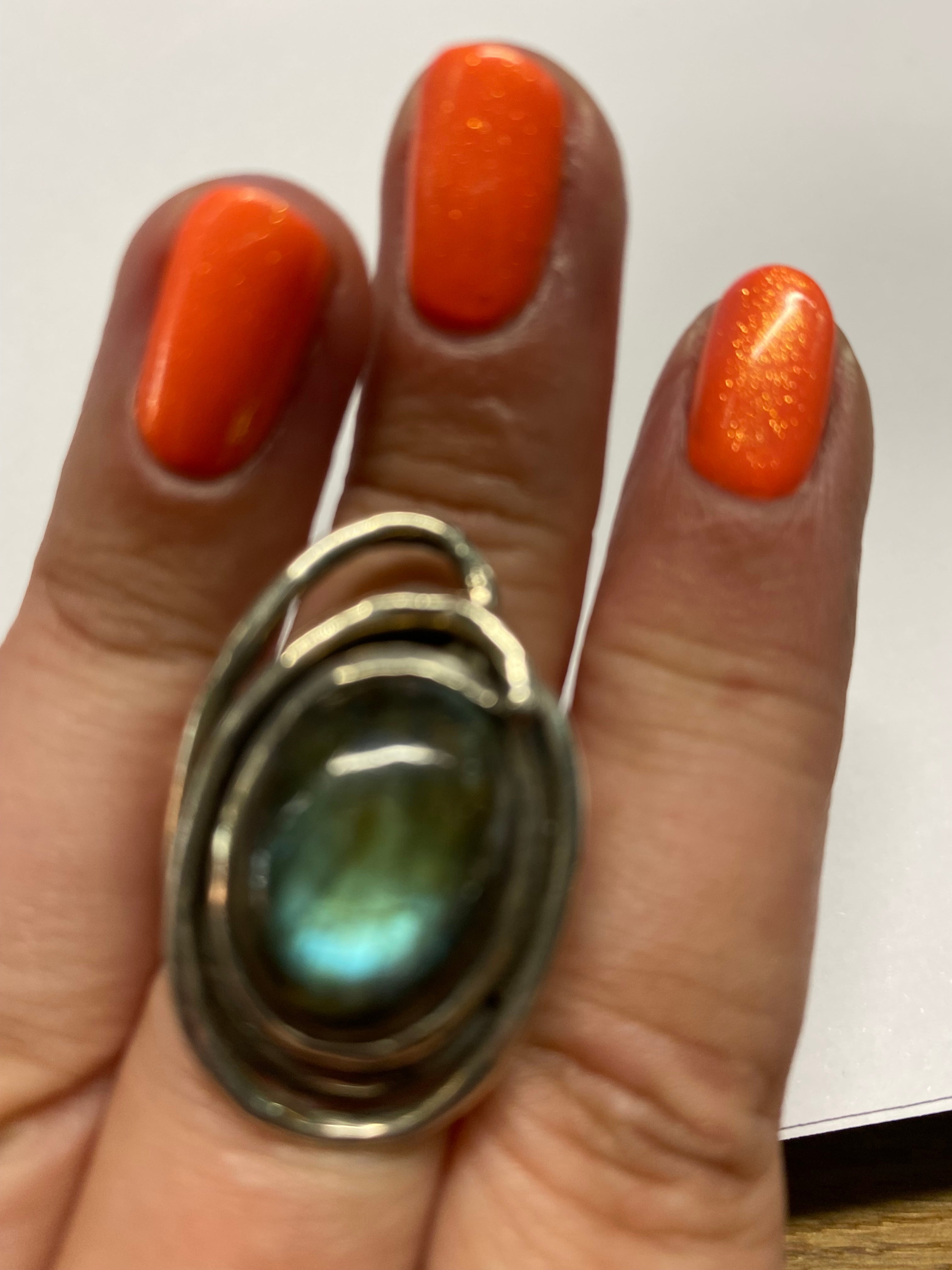 A hand with brightly painted orange nails is shown wearing a large, oval-shaped gemstone ring featuring a reflective, iridescent blue-green stone, set in a silver band with an intricate design. The background is blurry, focusing attention on the stunning 