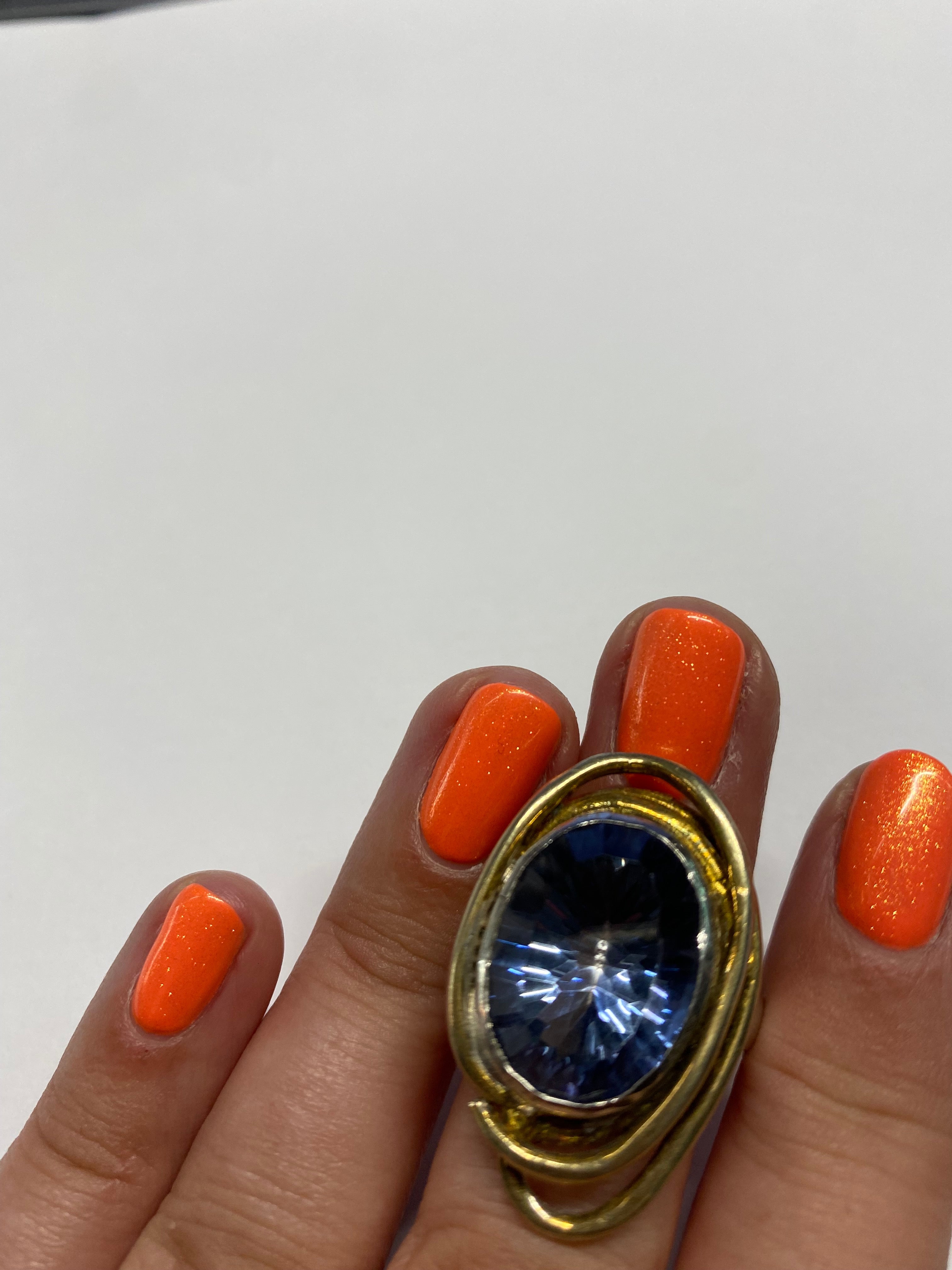 A hand with bright orange nail polish is shown against a plain background. The hand is wearing a large gold ring featuring a prominent, oval-shaped blue gemstone. The Cosmos Ring Large in Various Stones by Gallardo & Blaine Designs features gold-plated ac