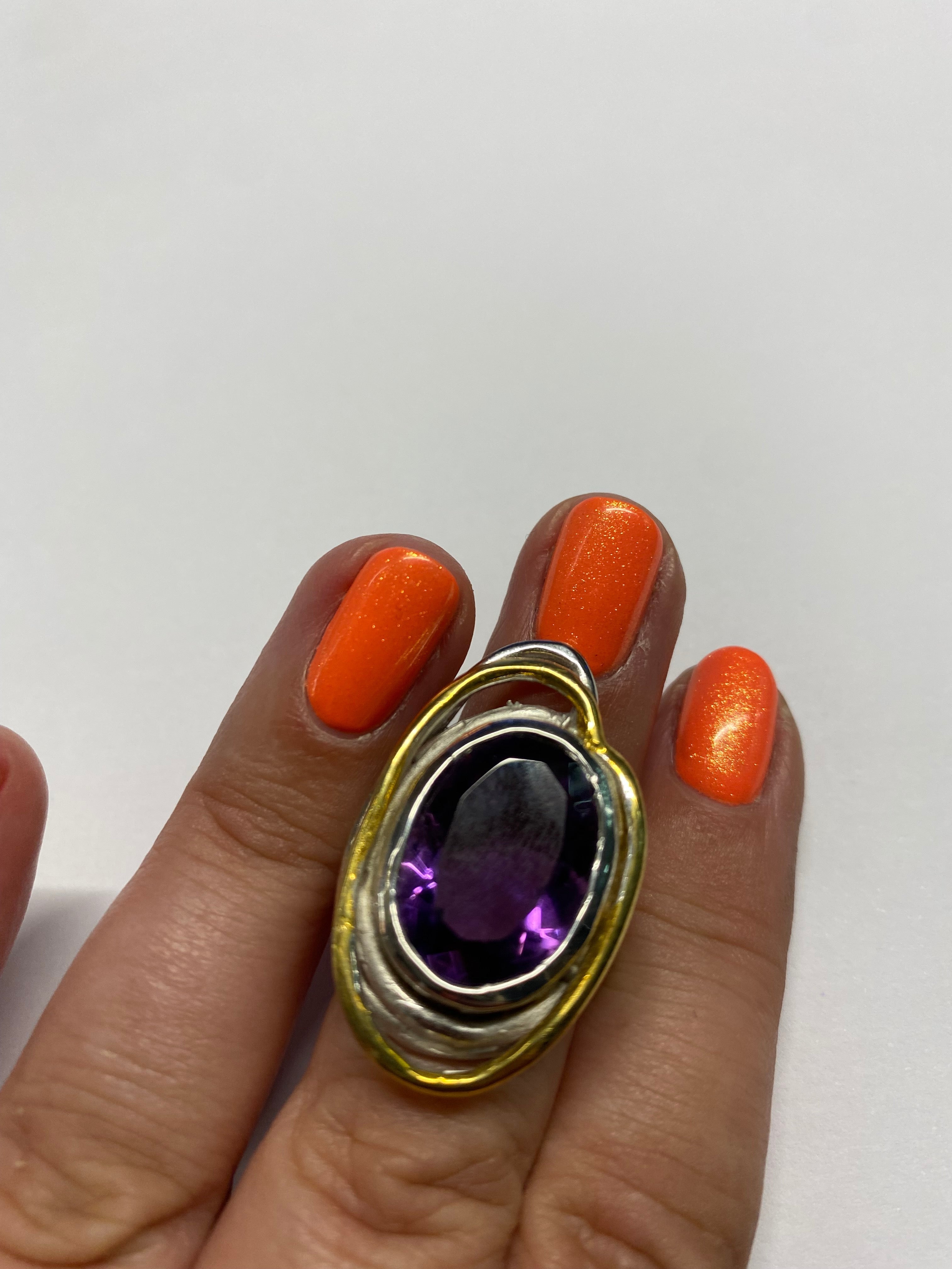 A hand with bright orange, glittery nail polish is shown. The middle finger is adorned with the Cosmos Ring Large in Various Stones by Gallardo & Blaine Designs, featuring a central purple gemstone, surrounded by metal layers in gold and silver tones. The