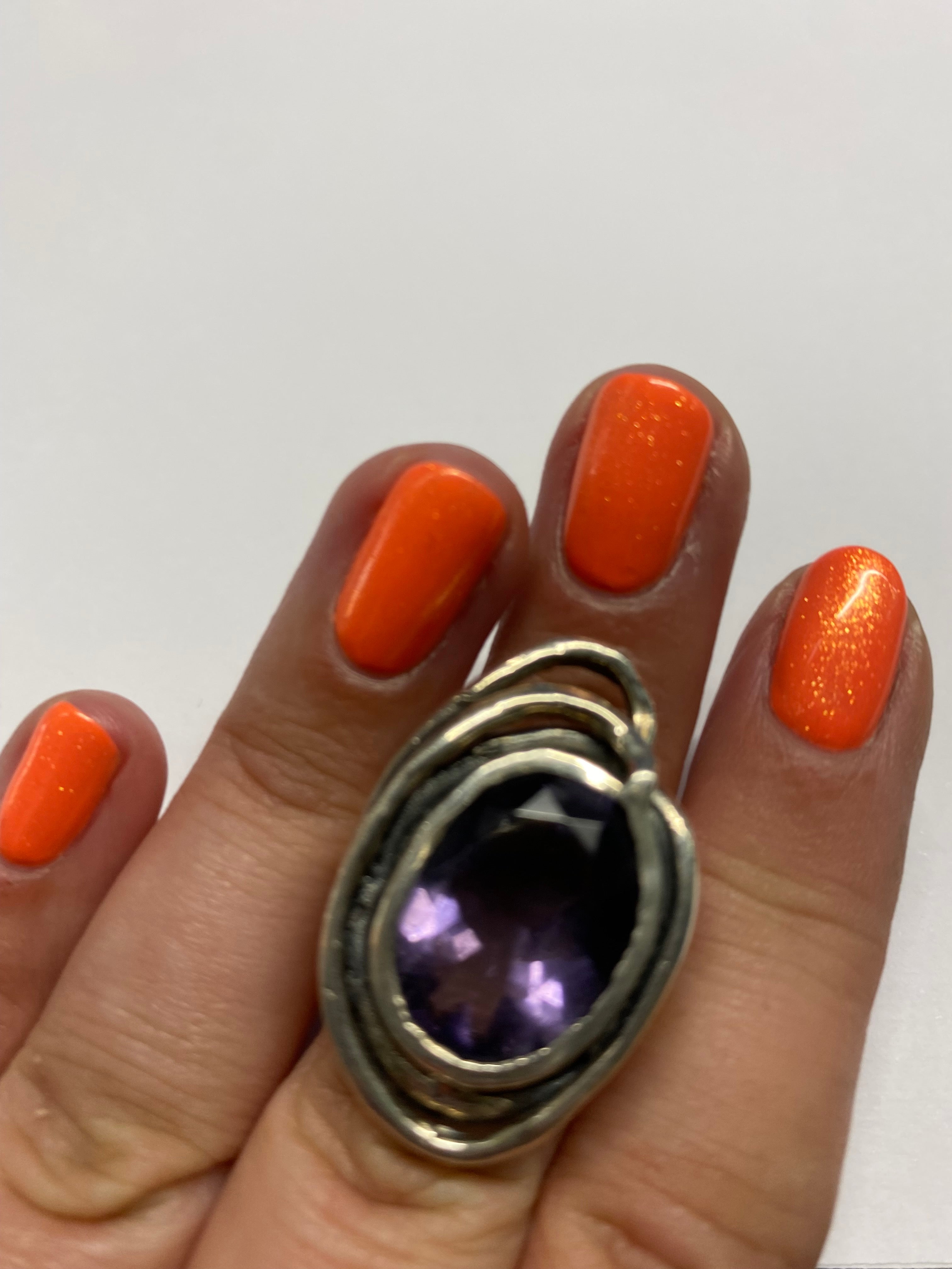 A hand with bright orange, glittery nail polish holds a silver ring featuring a large, dark purple gemstone from Gallardo & Blaine Designs' Cosmos Ring Large in Various Stones collection. The background is a plain white surface, accentuating the striking 