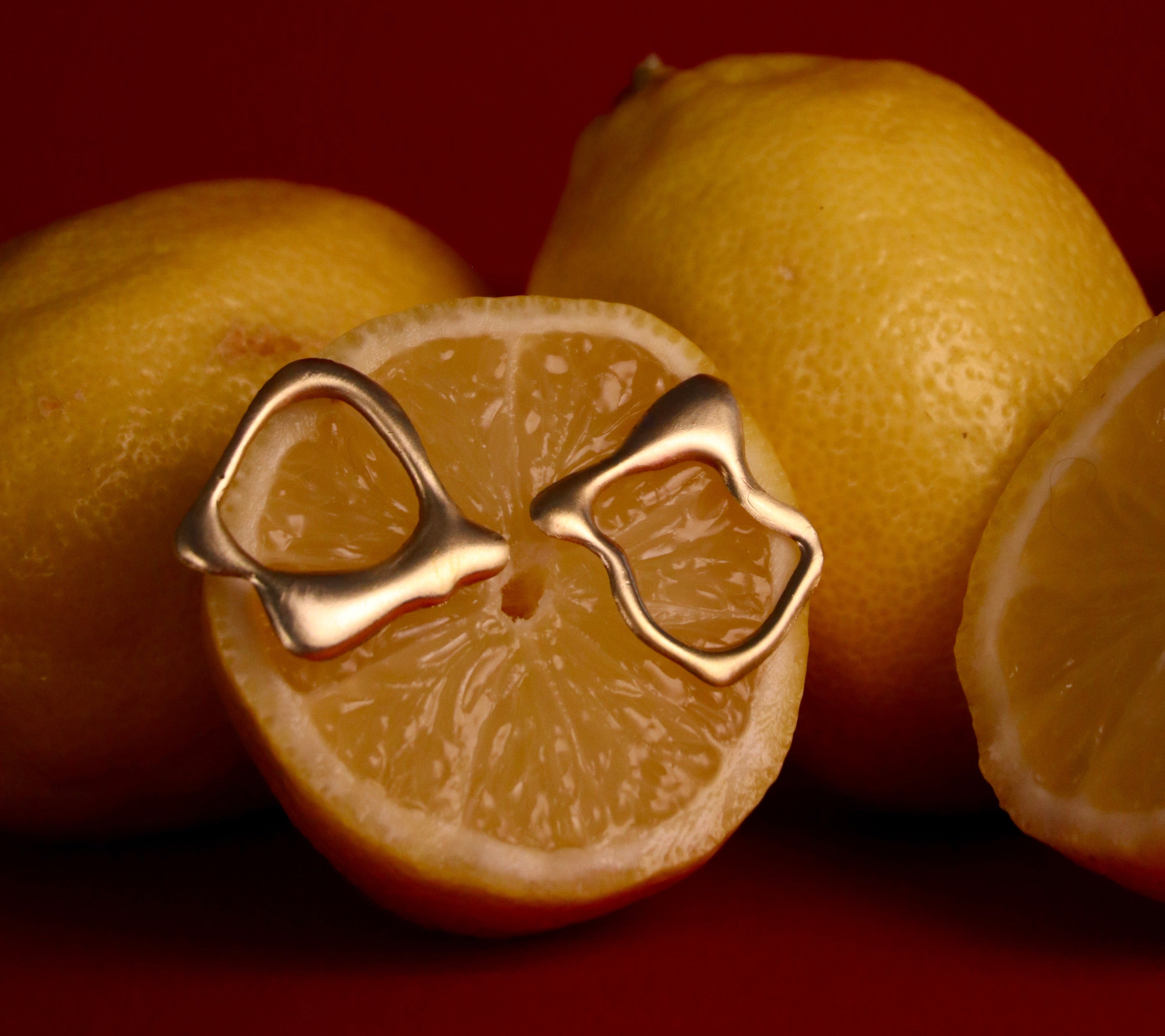 Three lemons rest on a red surface, one sliced in half. Two Asymmetrical Molten Studs by Madeleine Sculptural Jewellery are displayed on the juicy flesh of the halved lemon.