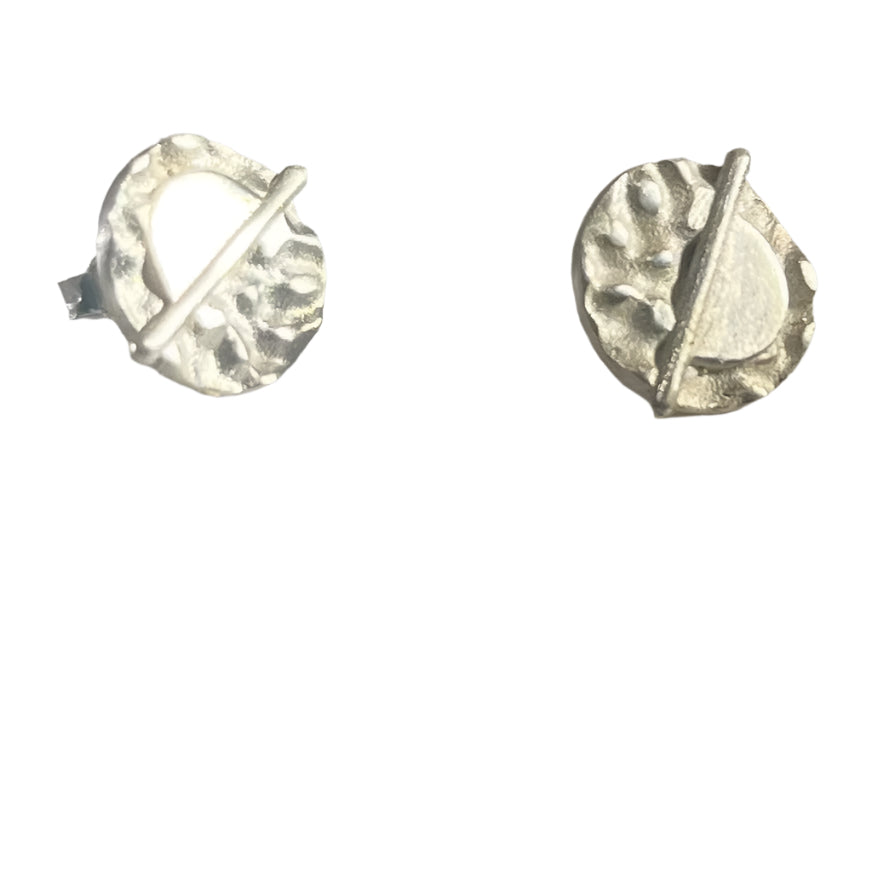 A pair of Lunar Eclipse Stud Earrings from Gallardo & Blaine Designs on a light surface. Each earring features an irregularly textured round design with an angled bar crossing the circle, creating a modern, abstract appearance reminiscent of Art Deco. The