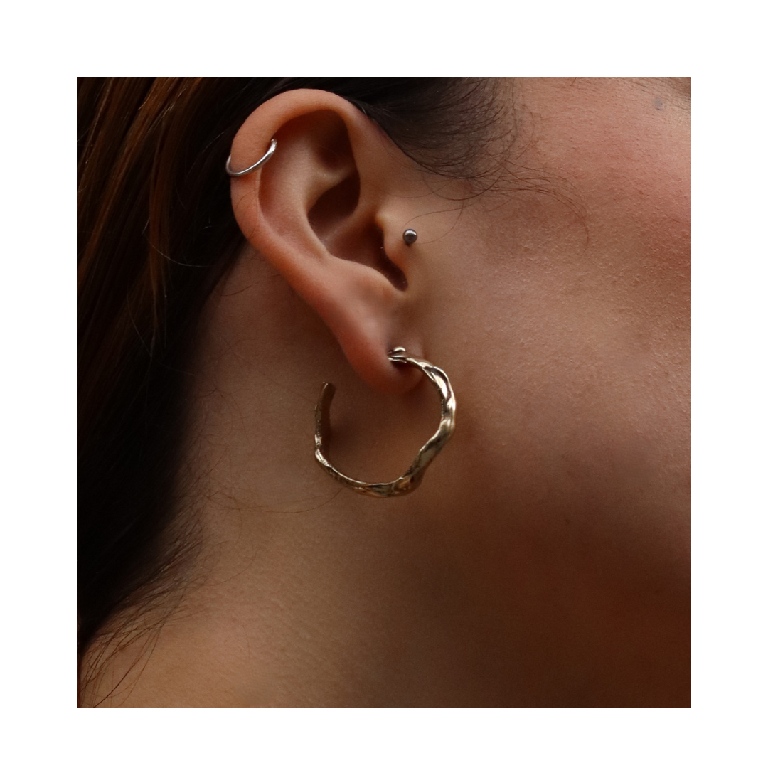 Close-up of a person's ear adorned with three earrings: a small hoop on the upper ear, a stud in the middle, and the Liana Hoop—a large, hand-carved gold earring from Madeleine Sculptural Jewellery—on the lower lobe. The plain white background highlights 