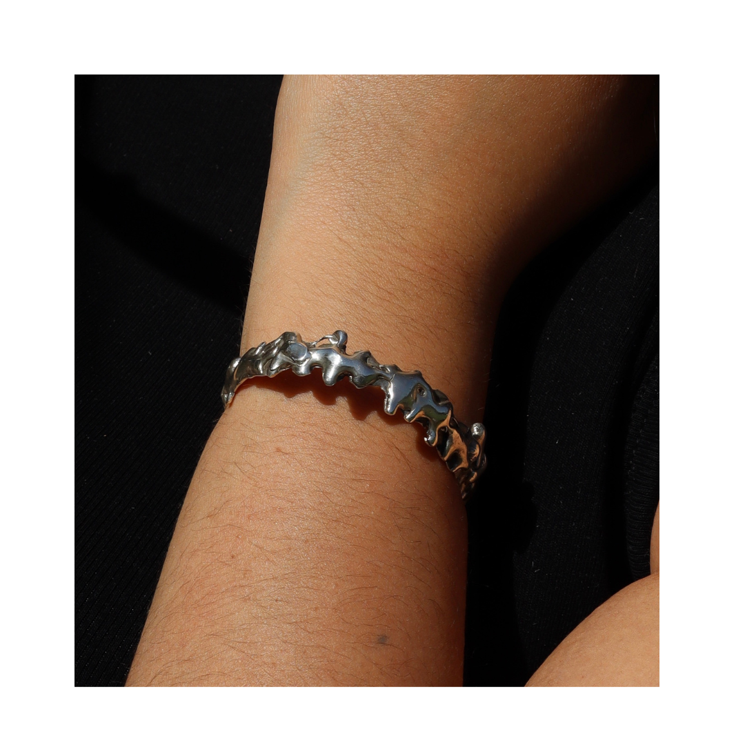 A close-up of a wrist adorned with the Molten Cuff Small from Madeleine Sculptural Jewellery, highlighting its shiny, irregular silver design and handmade elegance against a black fabric background.