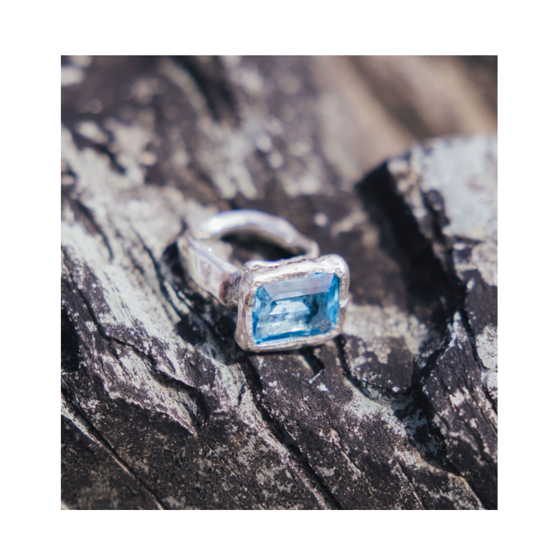 The Glacier Ring by Madeleine Sculptural Jewellery, featuring a rectangular blue gemstone set in silver, rests on a textured, dark stone surface.