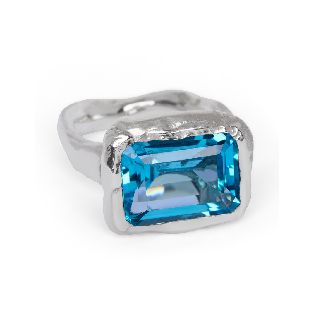 The Glacier Ring by Madeleine Sculptural Jewellery features an irregular, organic silver band set with a large rectangular blue gemstone, displayed against a white background.