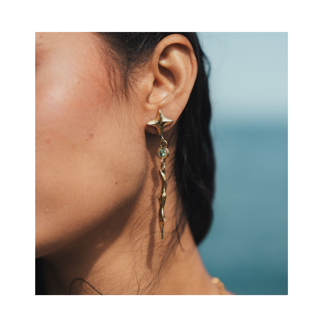 A close-up of a woman’s ear wearing Madeleine Sculptural Jewellery’s Nova Earrings Small with Moissanite—a long, gold, twisted design featuring a sparkling moissanite stone. The background is blurred, revealing blue sky and water.
