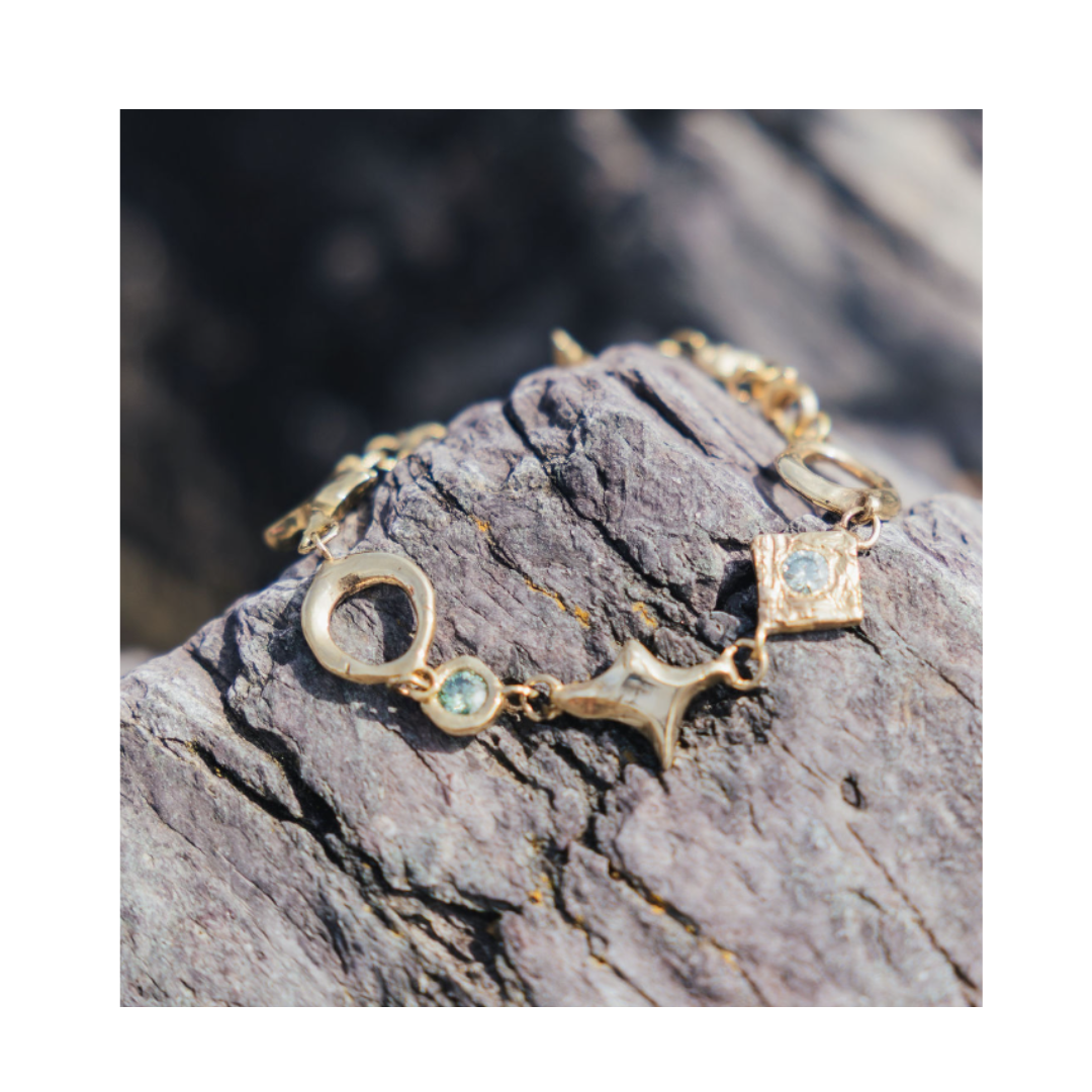 The Spark Bracelet in Brass by Madeleine Sculptural Jewellery, featuring distinctive geometric charms and small blue gemstones, rests on a textured gray rock in natural light.
