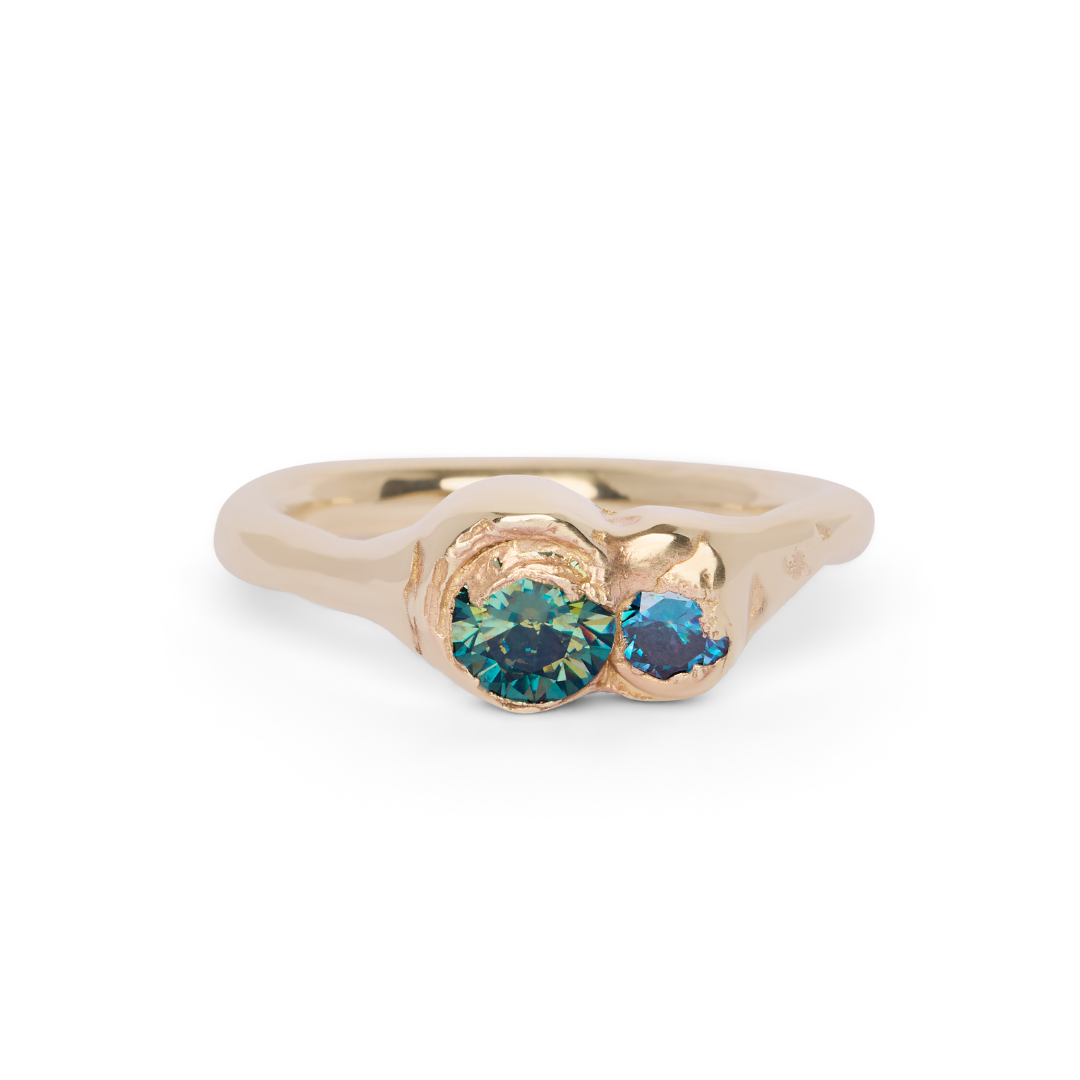 The Dune Ring by Madeleine Sculptural Jewellery features an organic, wavy gold band with two round, blue-green gemstones set side by side in an asymmetrical bezel-style setting.
