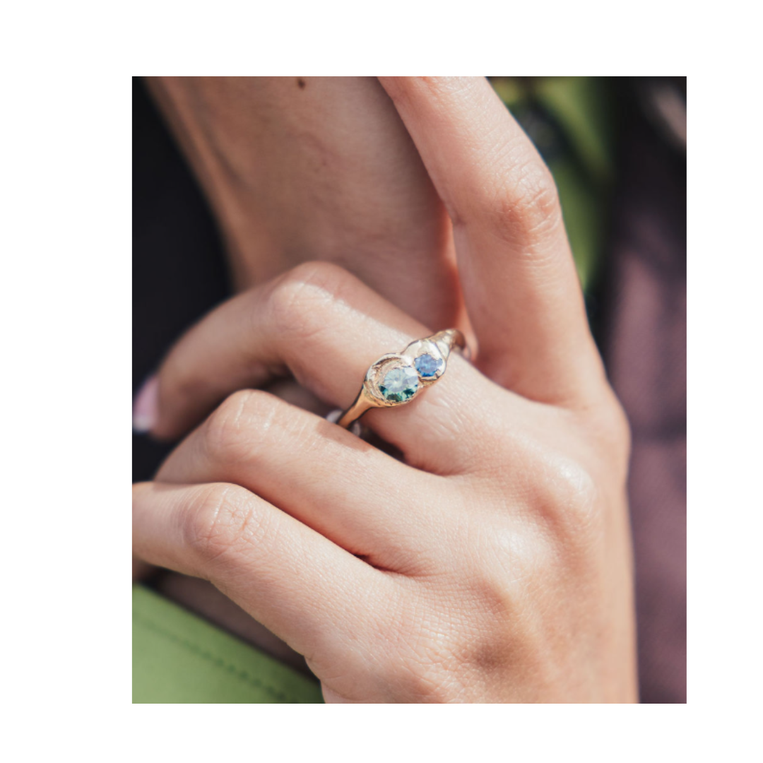 A close-up of a hand wearing the Dune Ring by Madeleine Sculptural Jewellery, featuring three stones—two clear and one green—set in gold on the ring finger, with the fingers slightly bent and a blurred background.