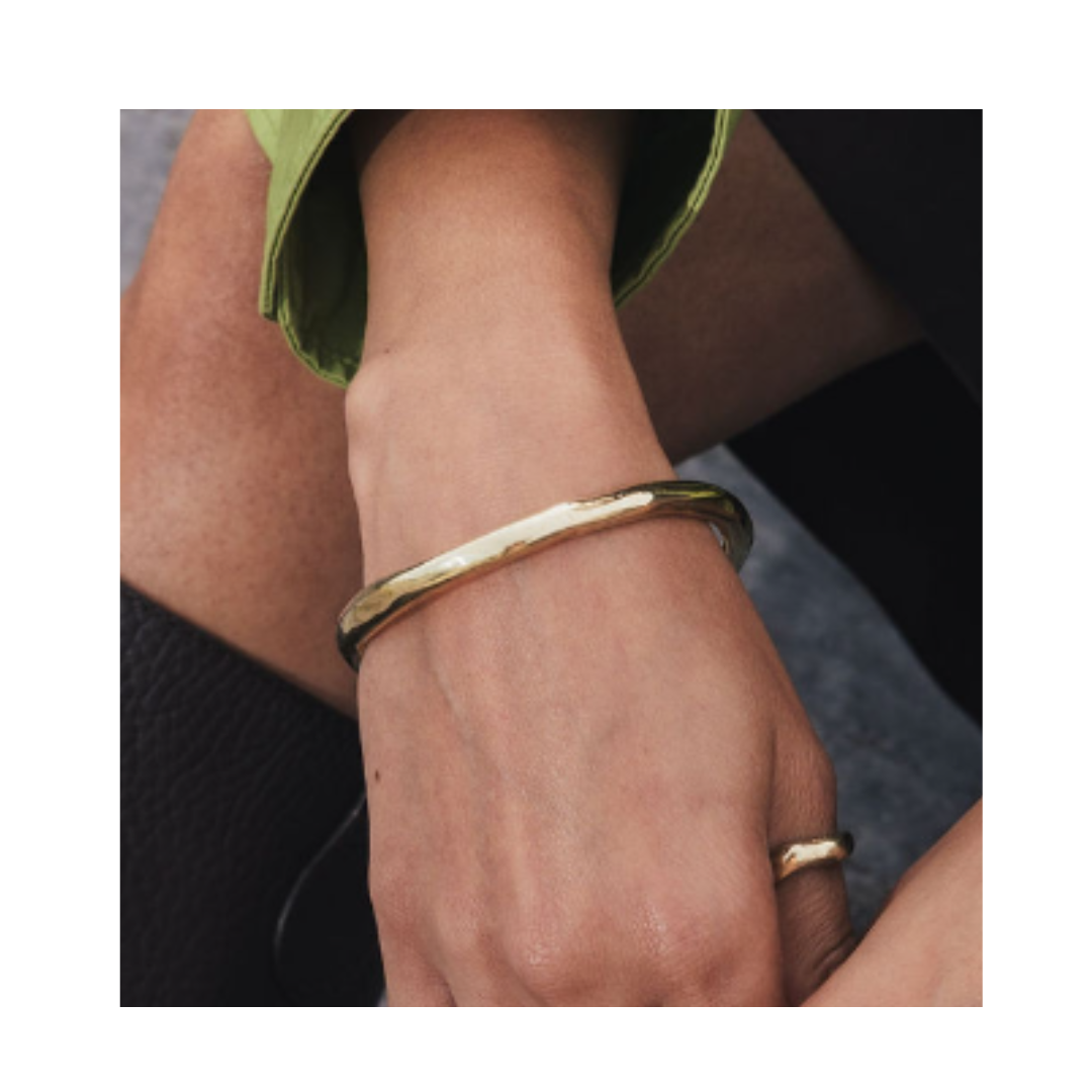 A close-up of a person’s hand wearing the Thorn Bangle by Madeleine Sculptural Jewellery and a gold ring. The person has a light brown skin tone and is dressed in a green sleeve and black pants.