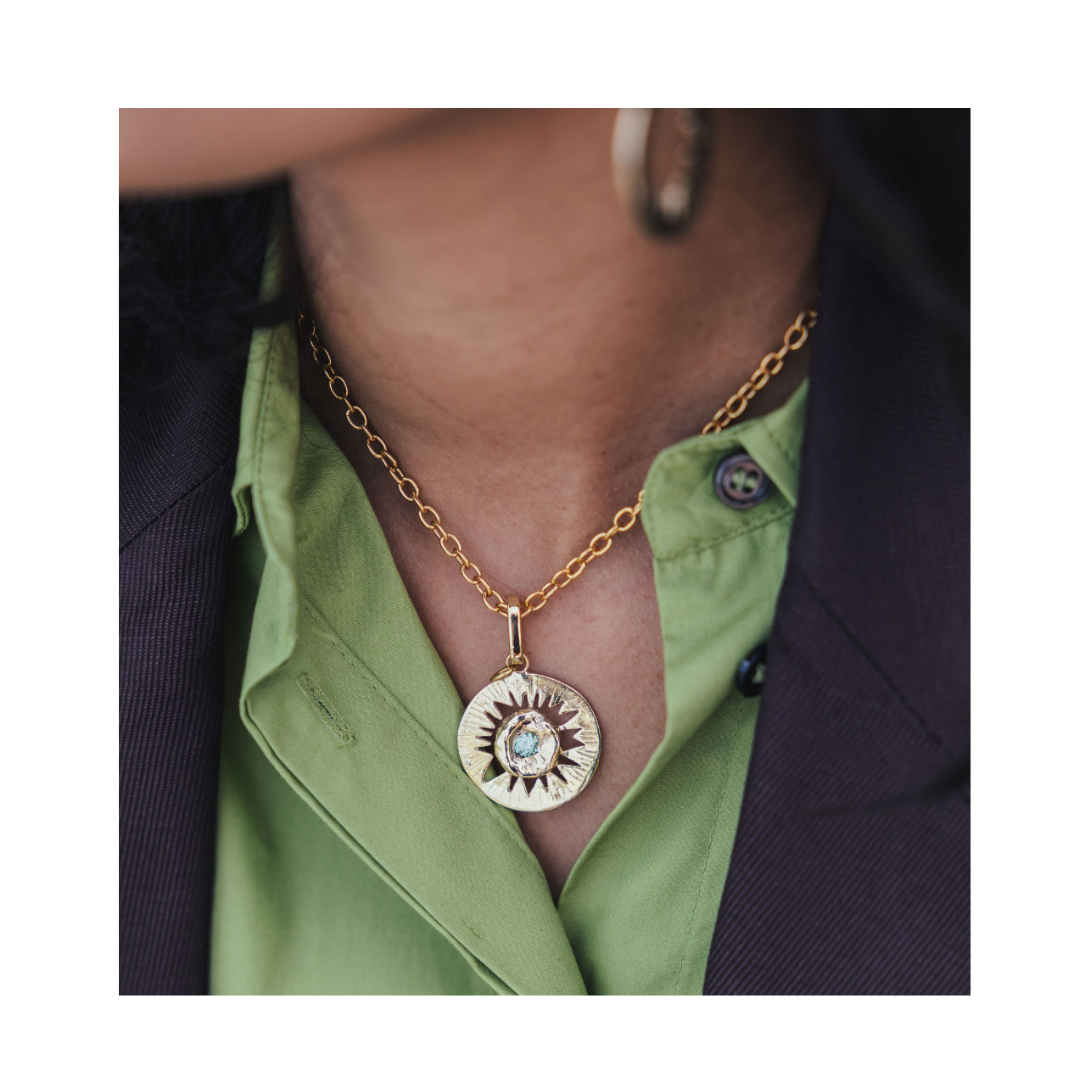 A person wearing the Tundra Necklace by Madeleine Sculptural Jewellery—a gold chain necklace with a large, round sunburst pendant—paired with a green button-up shirt under a dark blazer.
