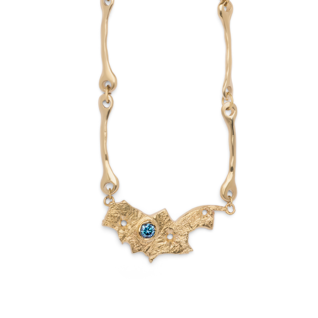 The Blaze Necklace by Madeleine Sculptural Jewellery features a bat-shaped pendant with a textured surface, adorned with small pearl accents and a striking central blue gemstone. Its chain is designed with organic, wavy links. Photographed on a white back