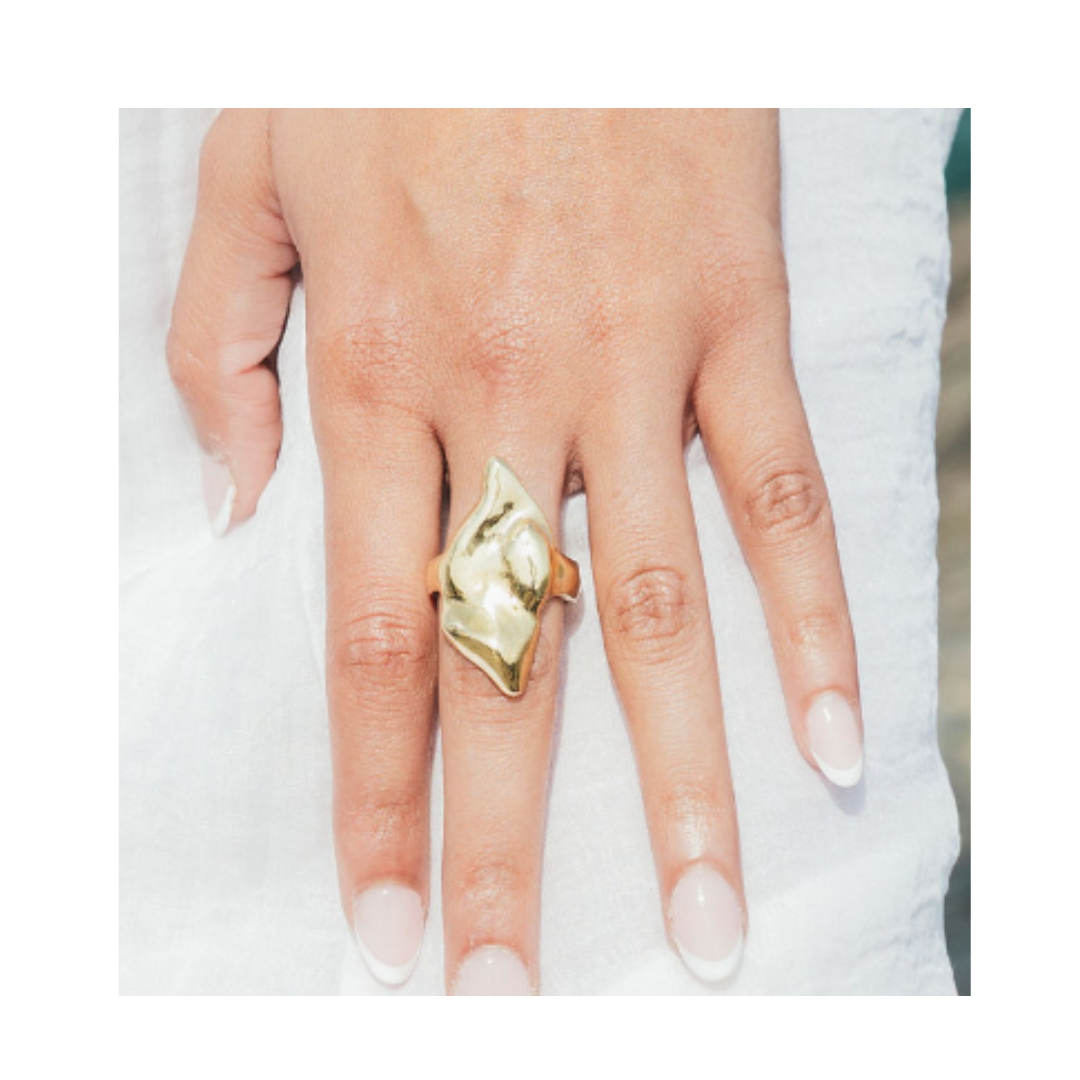 A hand with neatly manicured nails wears the Rift Ring by Madeleine Sculptural Jewellery, a large abstract gold statement piece, on the middle finger. The background is white fabric.
