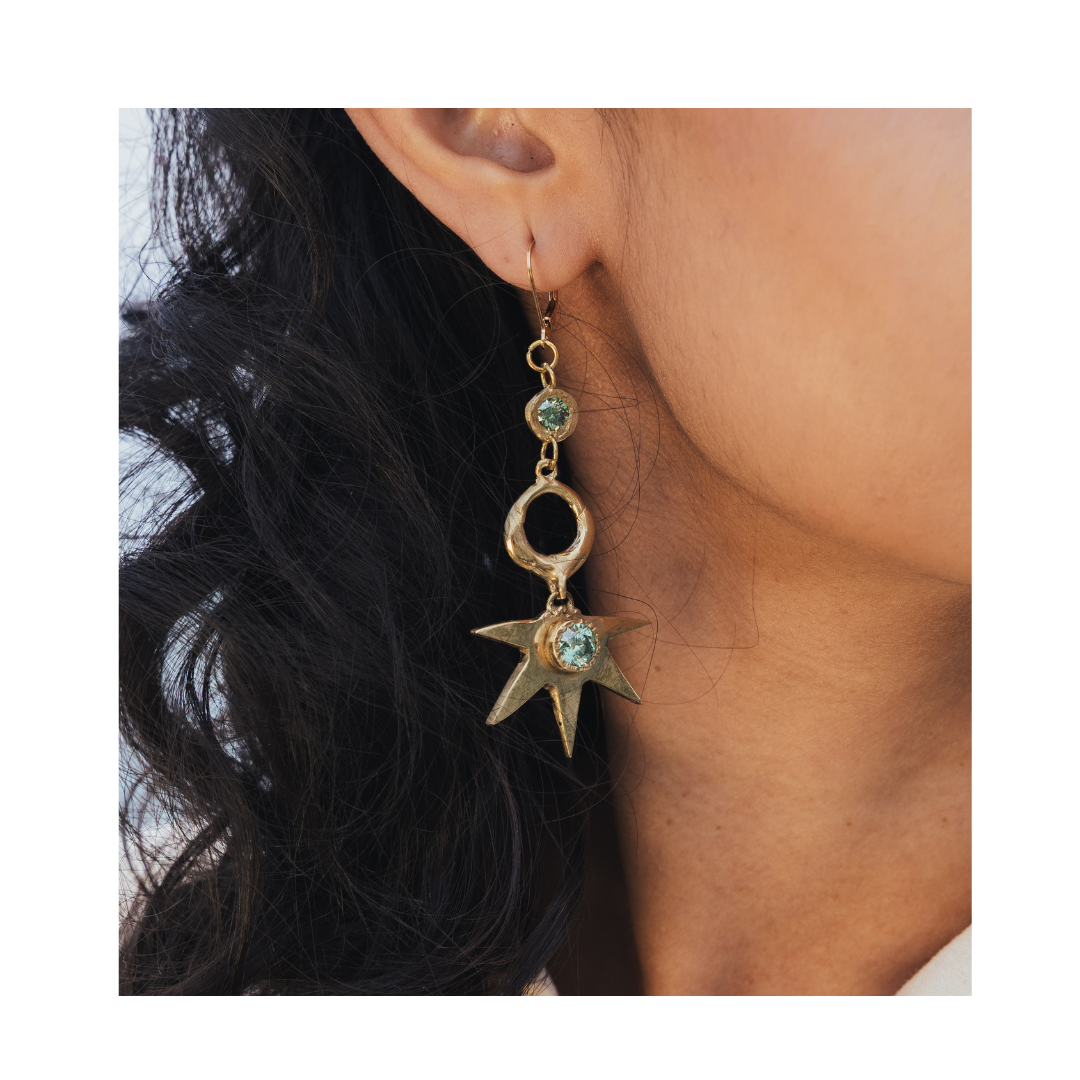 A close-up of a person with long, wavy dark hair wearing the Réalta Earrings with 2 moissanite by Madeleine Sculptural Jewellery—a striking 3-part gold earring adorned with a moon and star design, featuring a round gem at the top and a sparkling star-shap