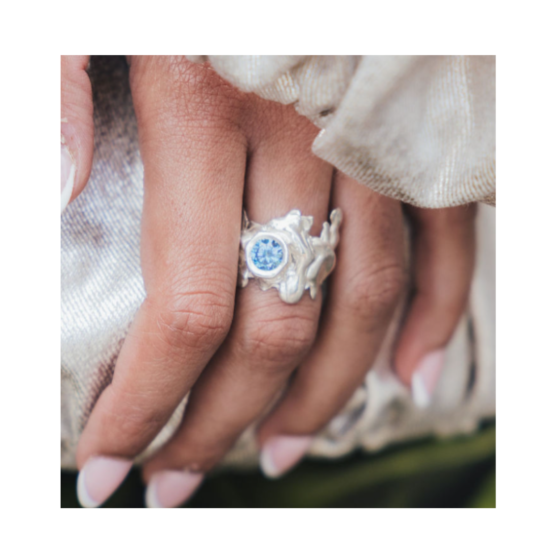 A close-up of a hand with manicured nails showcases the Solstice Ring with Large Moissanite by Madeleine Sculptural Jewellery. This striking example of custom design, set with a brilliant pale blue moissanite, rests elegantly on light, shimmery fabric—ref