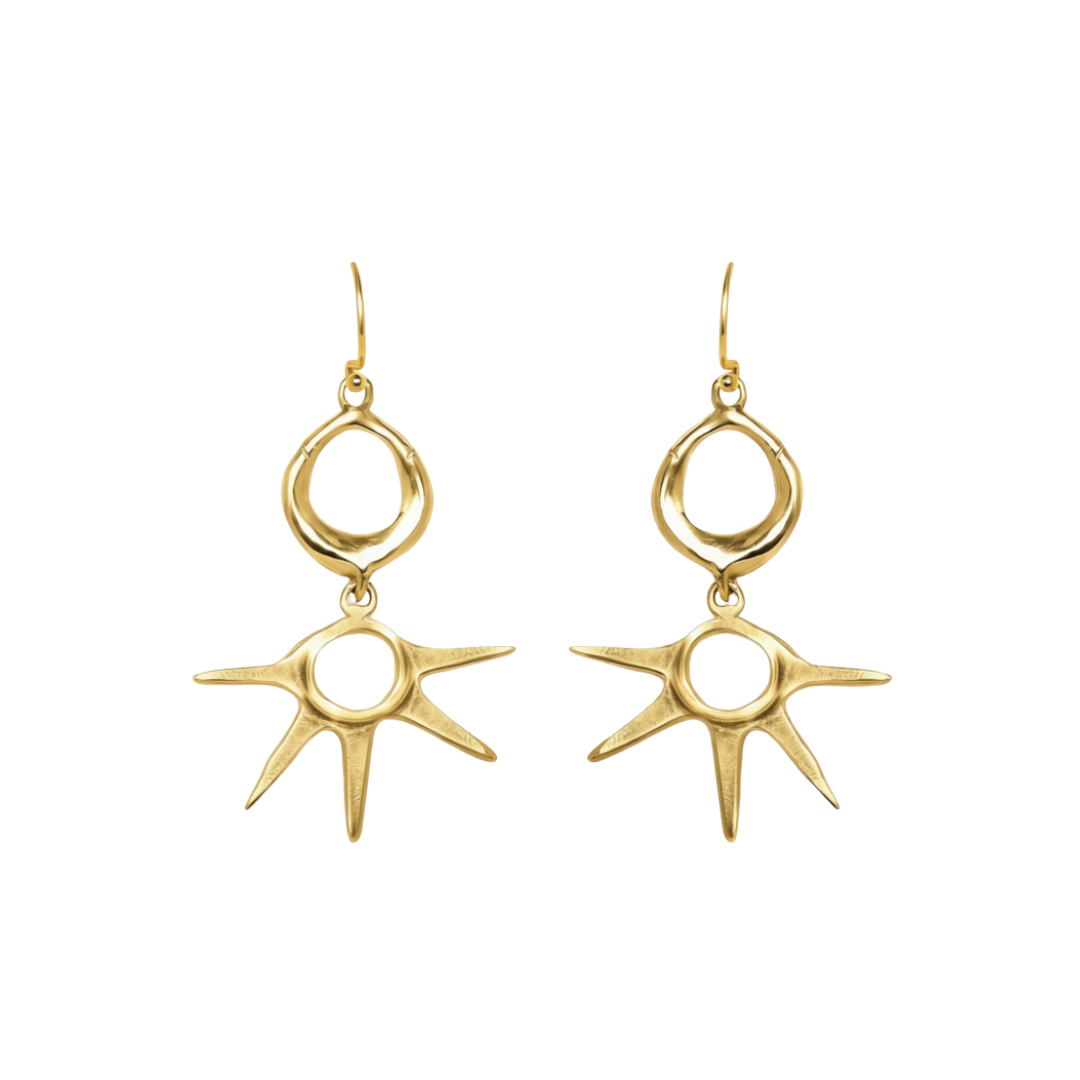 The Réalta Earrings by Madeleine Blaine are handcrafted Irish jewelry pieces, featuring gold dangle designs with an open oval at the top and a spiky sunburst motif below each centered around a circular hole. These luxury accessories are displayed against 