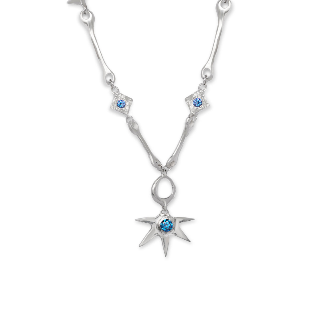 The Réalta Necklace with Moissanite by Madeleine Sculptural Jewellery features a central star-shaped pendant adorned with a blue moissanite gemstone, complemented by two smaller blue moissanites set in diamond-shaped accents along the silver chain.