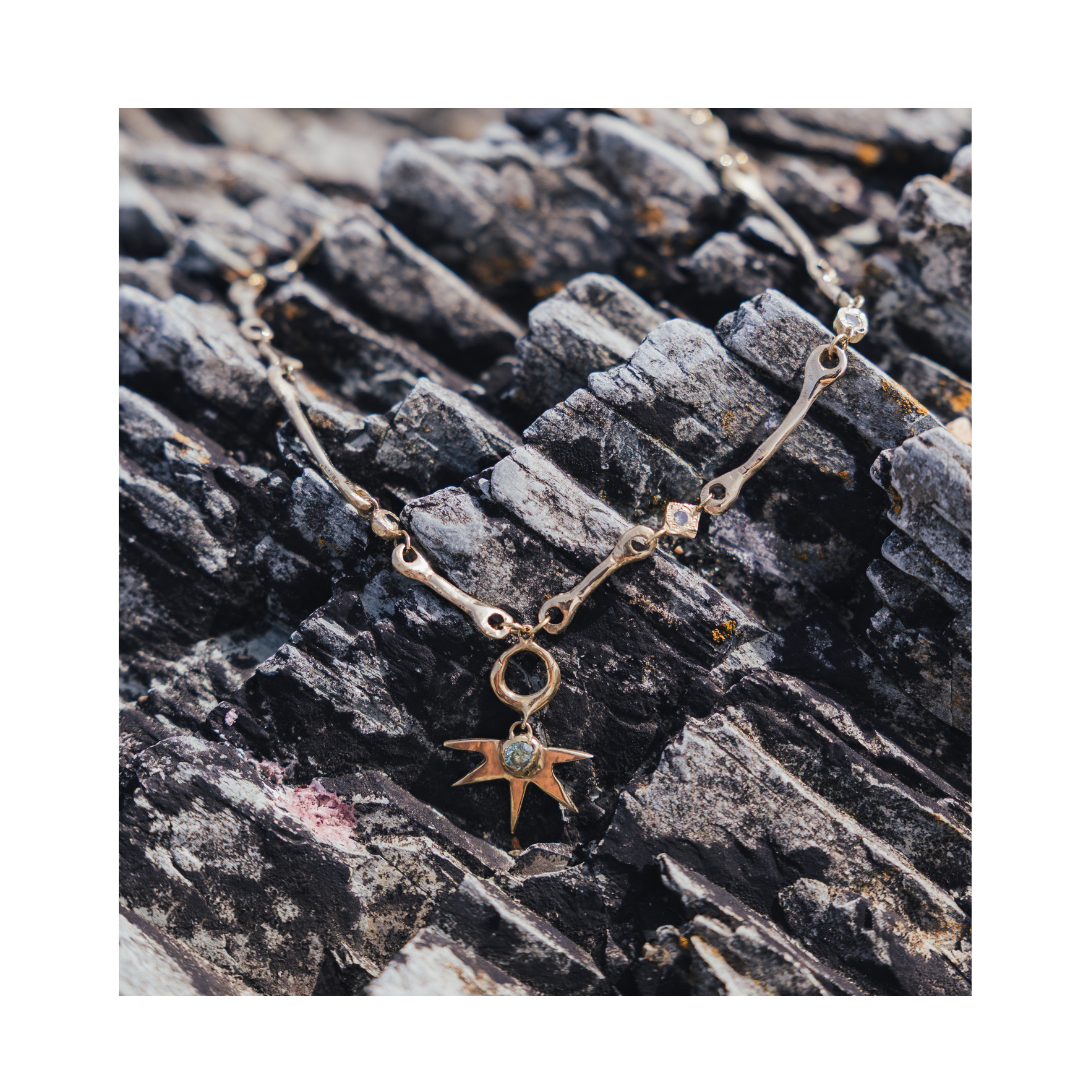 The Réalta Necklace with Moissanite by Madeleine Sculptural Jewellery, featuring a delicate gold chain adorned with sparkling moissanite stones and a star-shaped pendant, is elegantly displayed on textured dark gray rocks.