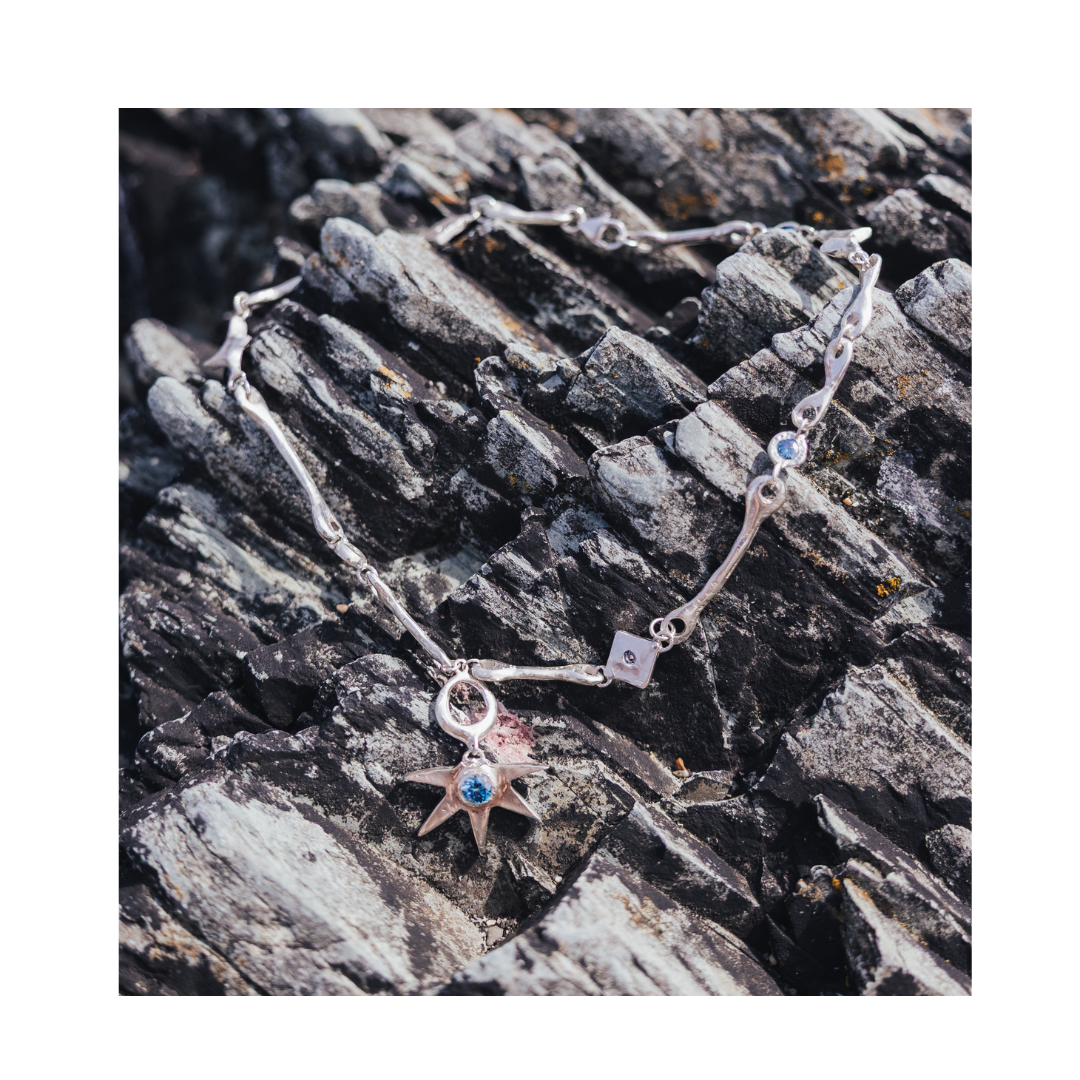 The Réalta Necklace with Moissanite by Madeleine Sculptural Jewellery, featuring a star-shaped pendant adorned with blue gemstones, is displayed on rough, layered gray rocks.