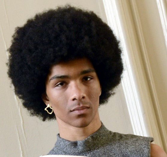 A person with a large, round afro hairstyle, wearing a sleeveless gray top and a gold heart-shaped earring, stands indoors against a light-colored wall. Their look is complemented by the elegant Asymmetrical Molten Studs from Madeleine Blaine, showcasing 