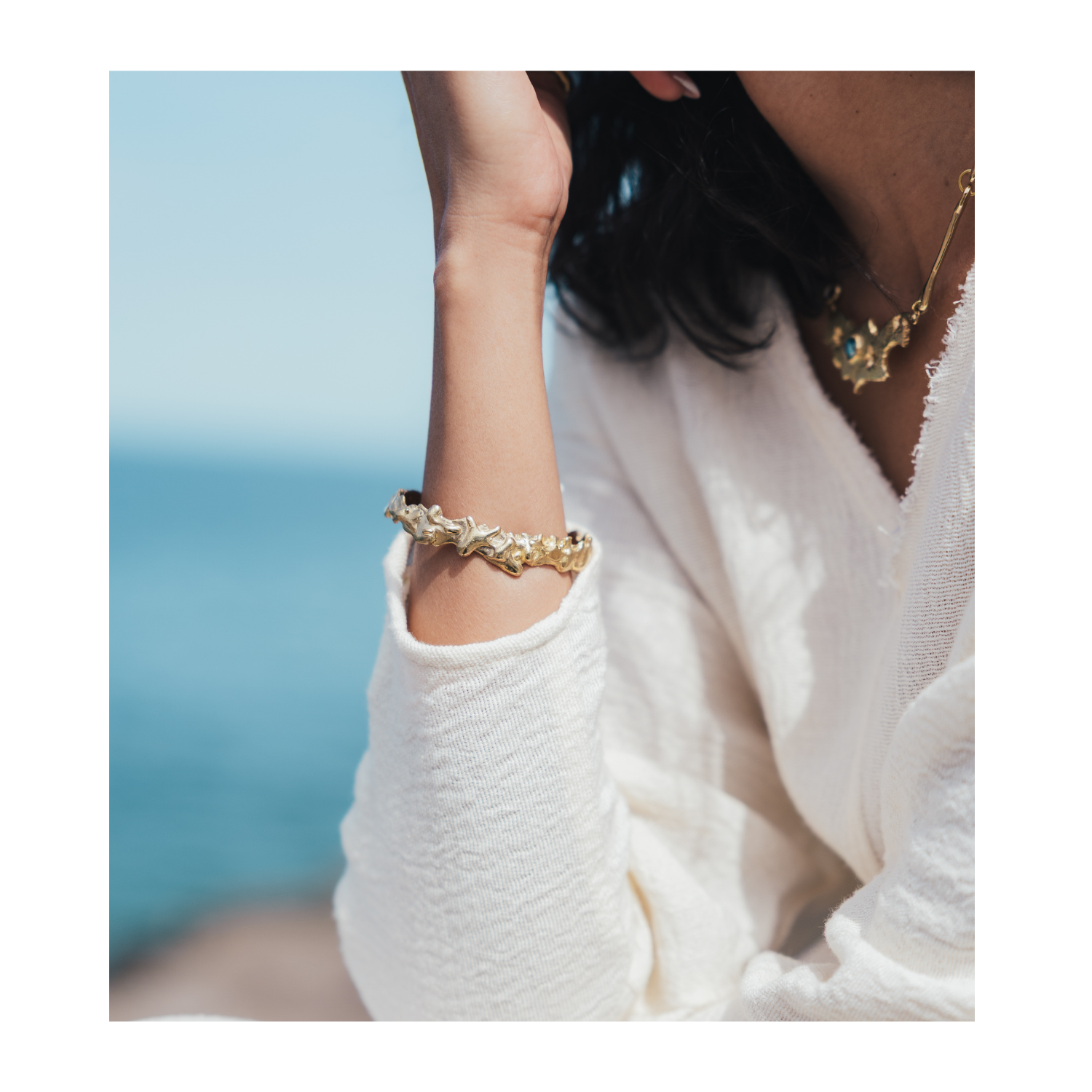 A close-up lifestyle shot of a person wearing a gold bracelet with clear crystal details on their wrist, paired with a white textured knit top and a gold necklace, with a blurred blue ocean background.