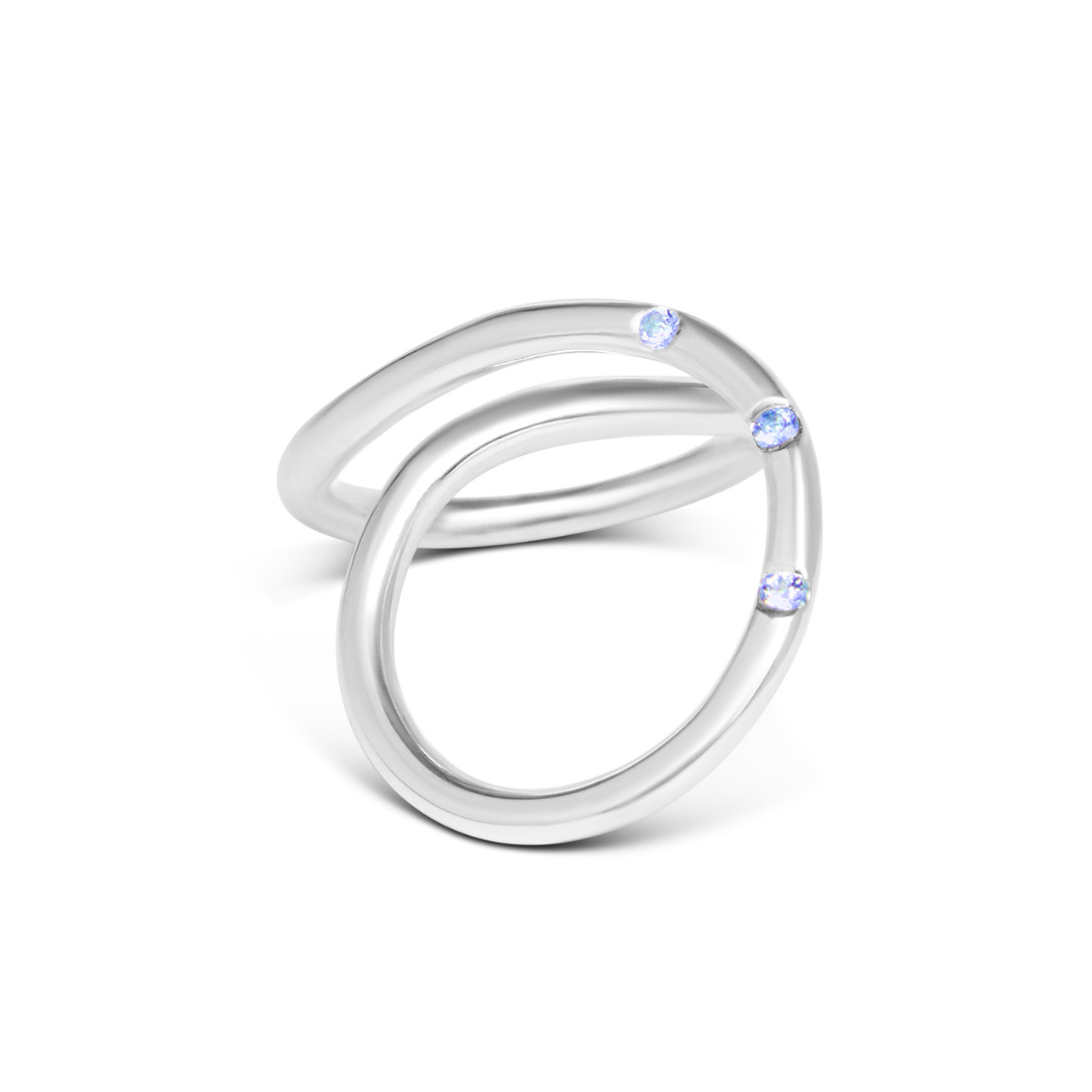 The Sea Honey Ring by Gallardo & Blaine Designs features two overlapping sterling silver loops, each set with a small round light blue gemstone, creating a minimalist and elegant look against a plain white background.