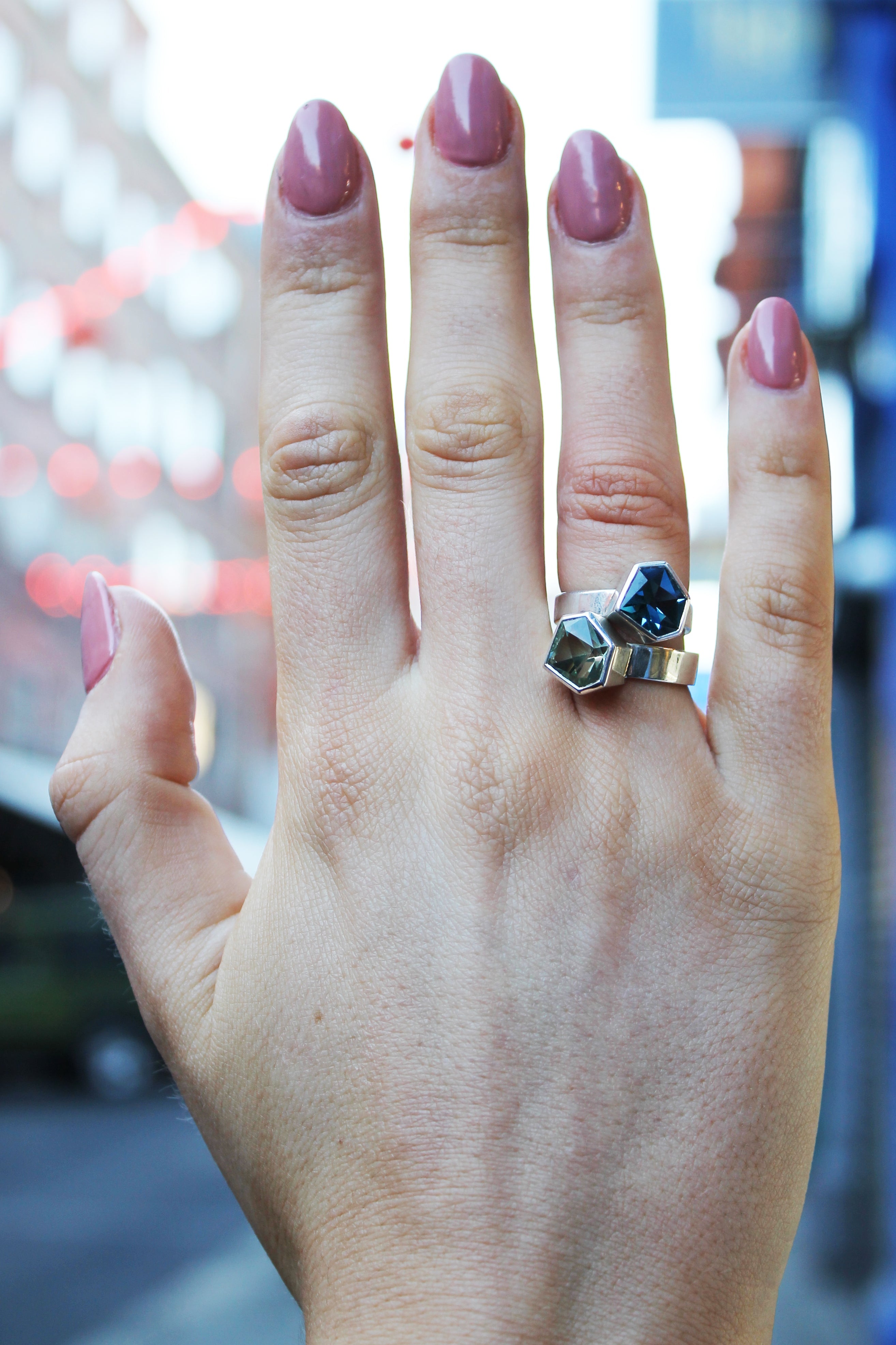 two rings, the first one, closer to the hand is a Silver ring with large, irregular hexagonal green amethyst in a silver bezel. Band is flat and 5mm wide.The second is a Silver ring with large, irregular hexagonal blue topaz in a silver bezel. Band is fla