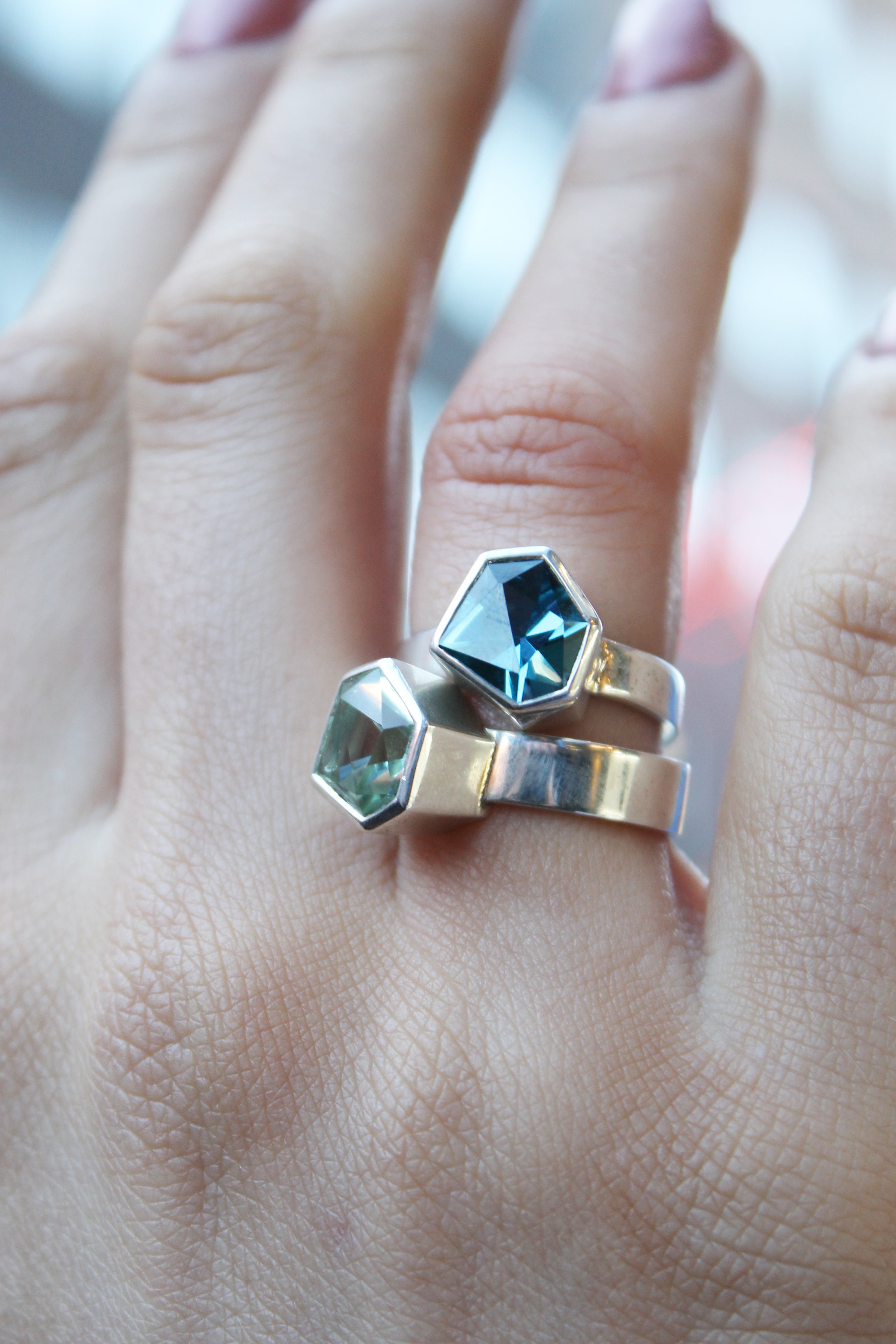 two rings, the first one, closer to the hand is a Silver ring with large, irregular hexagonal green amethyst in a silver bezel. Band is flat and 5mm wide.The second is a Silver ring with large, irregular hexagonal blue topaz in a silver bezel. Band is fla