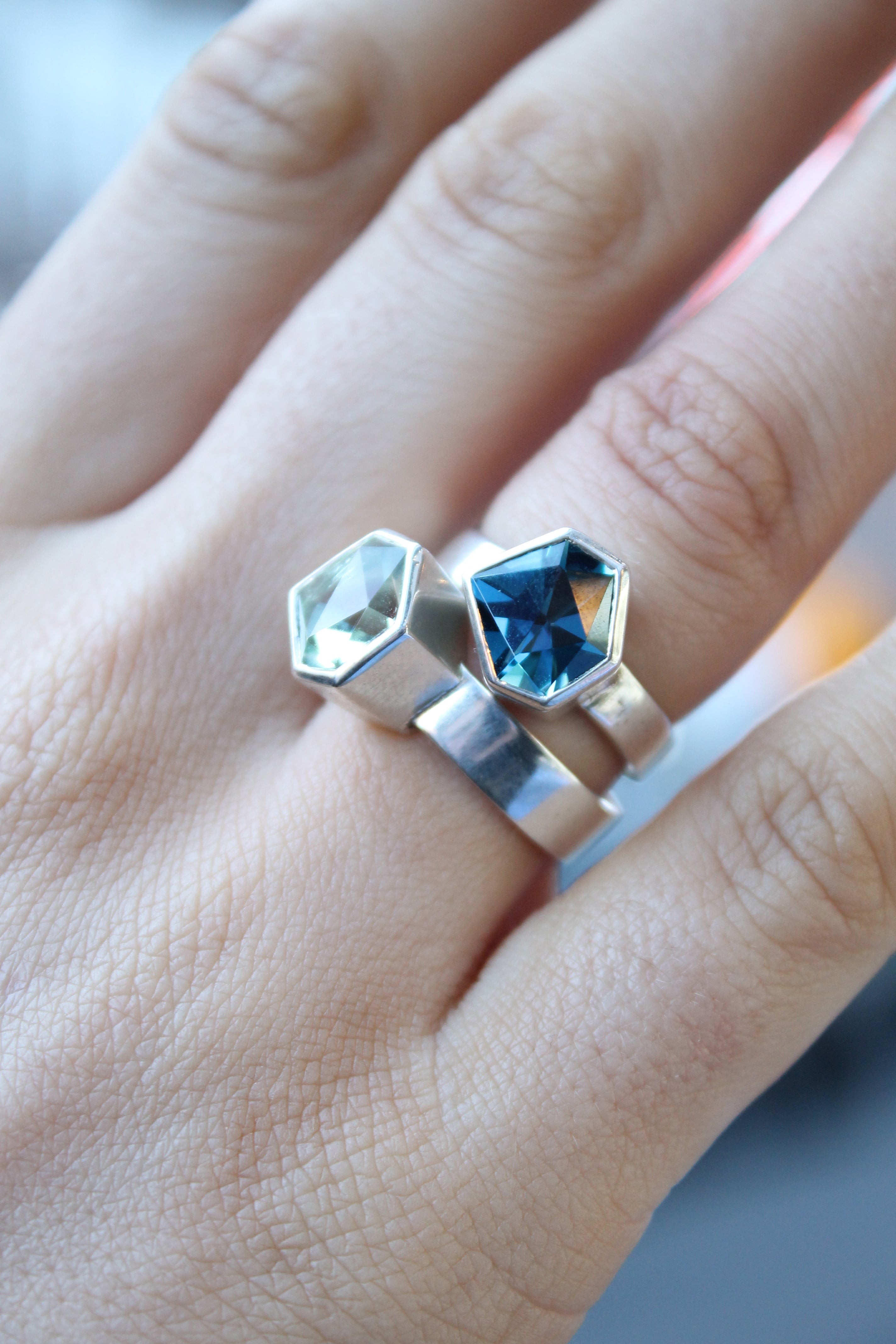 two rings, the first one, closer to the hand is a Silver ring with large, irregular hexagonal green amethyst in a silver bezel. Band is flat and 5mm wide.The second is a Silver ring with large, irregular hexagonal blue topaz in a silver bezel. Band is fla